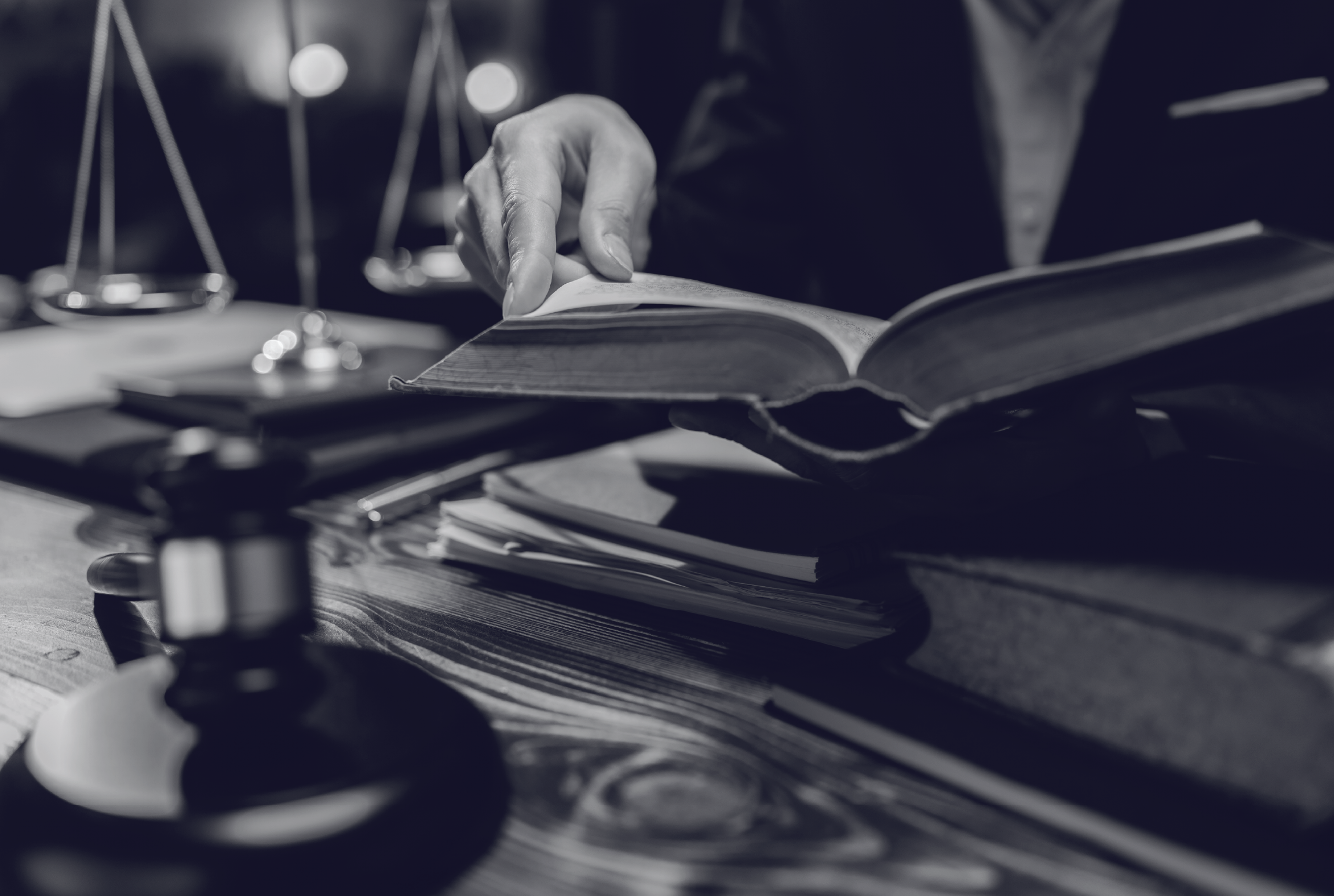 Una mano pasa las páginas de un libro en un escritorio con documentos y una balanza legal.