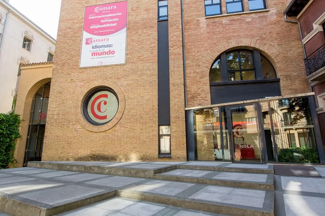 Edificio de ladrillos con cartel en la parte superior y una ventana circular con un logo en el centro, entrada principal con puertas de vidrio y escaleras de acceso.