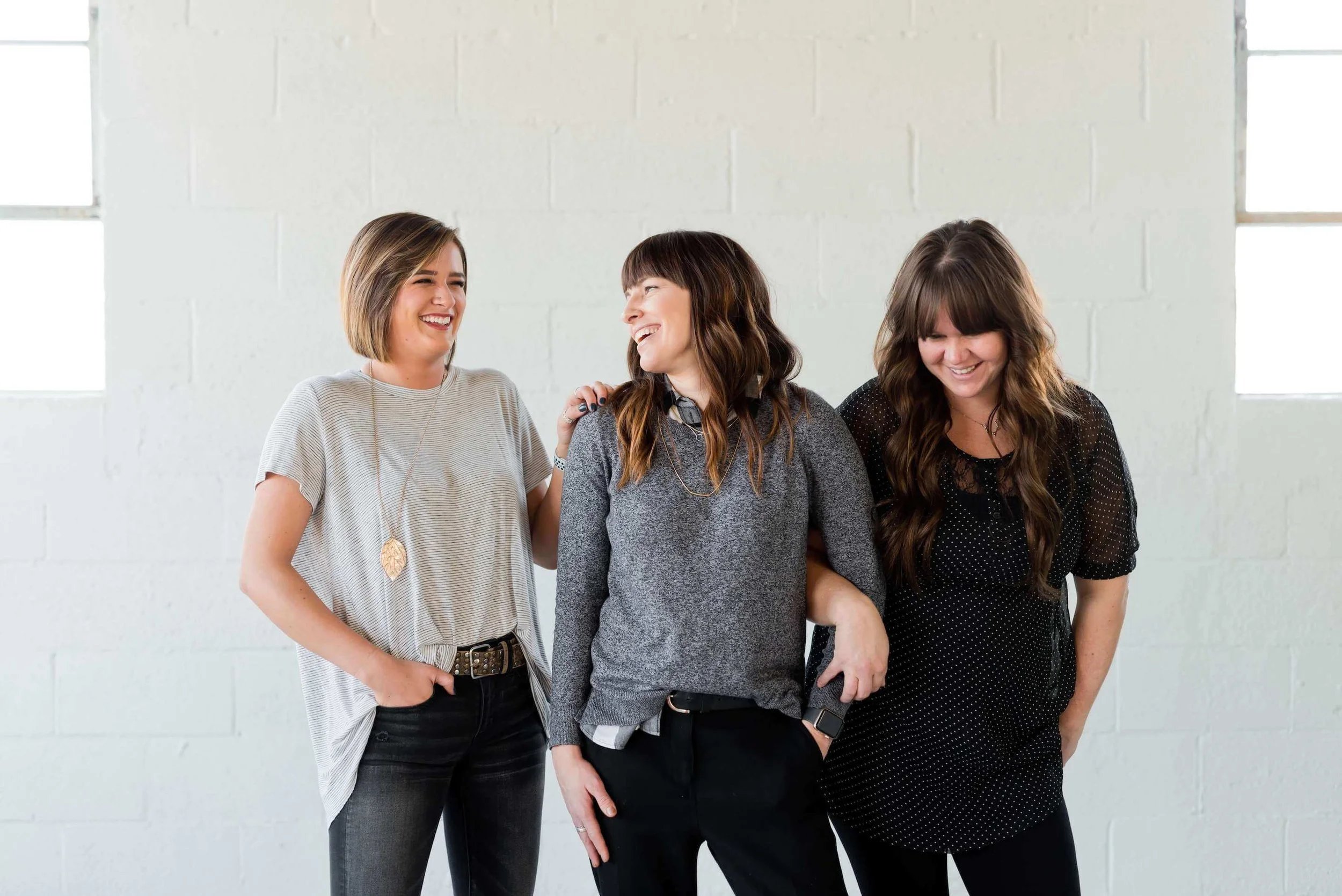 Three women smiling and laughing together, standing against a white brick wall. The woman on the left has short brown hair, wearing a light gray top and dark jeans. The woman in the middle has long brown hair with bangs, wearing a gray sweater and black pants. The woman on the right has long wavy brown hair with bangs, wearing a black dress with small white dots.