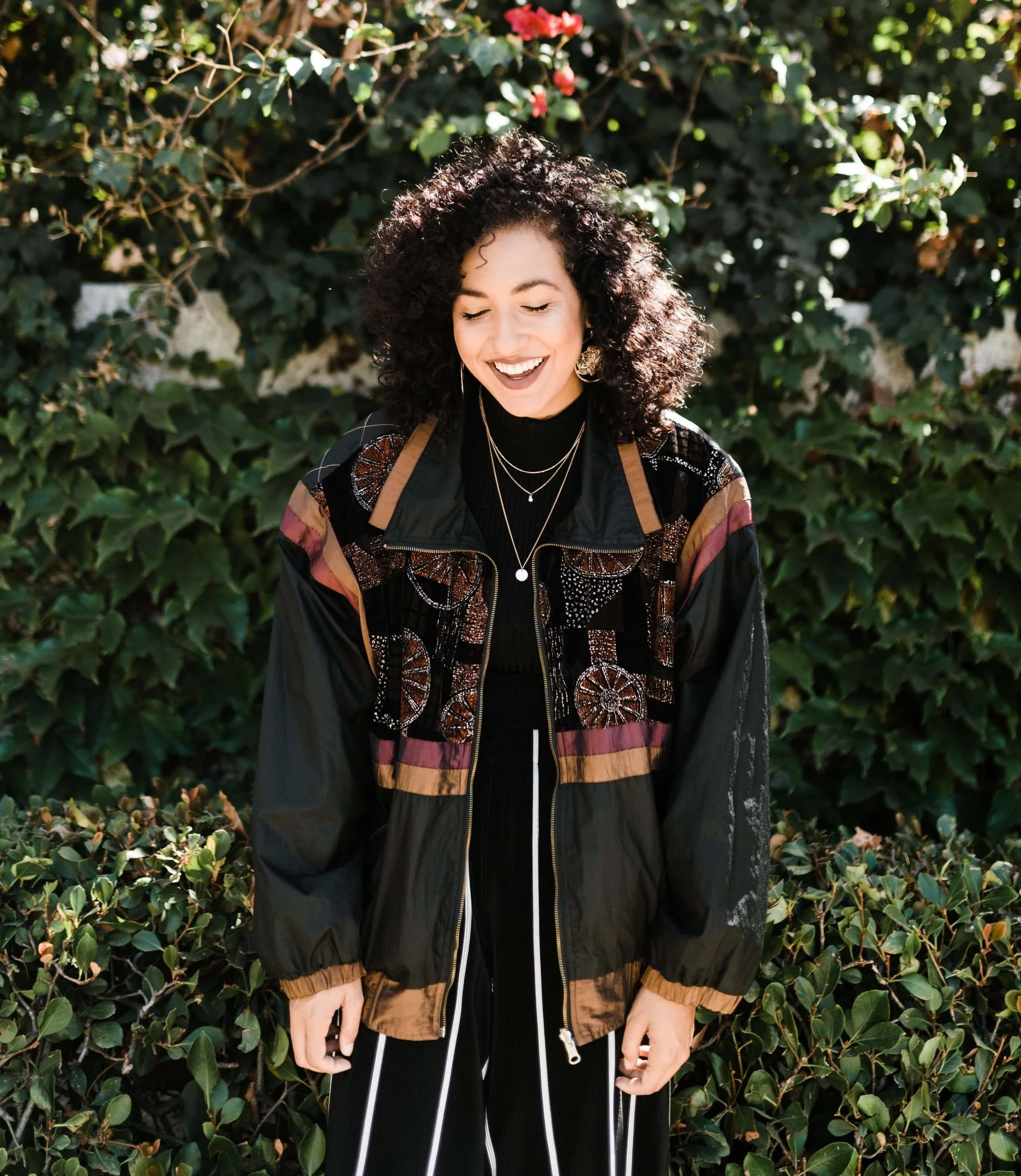 A woman with curly dark hair smiling and looking down, standing outdoors in front of green leafy bushes.