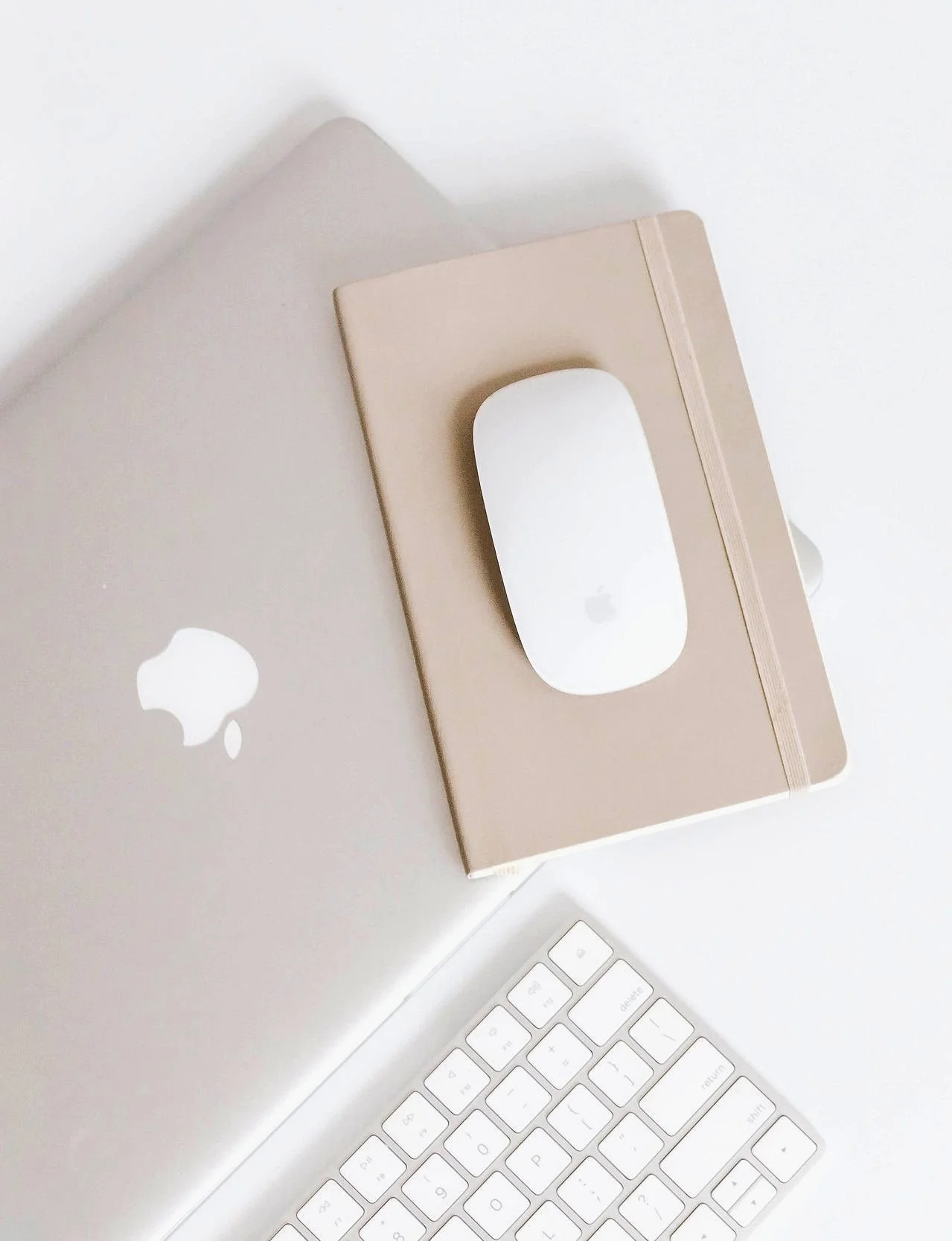 A closed silver laptop with an Apple logo, a beige notebook, a white Apple Magic Mouse, and a white Apple keyboard on a white surface.