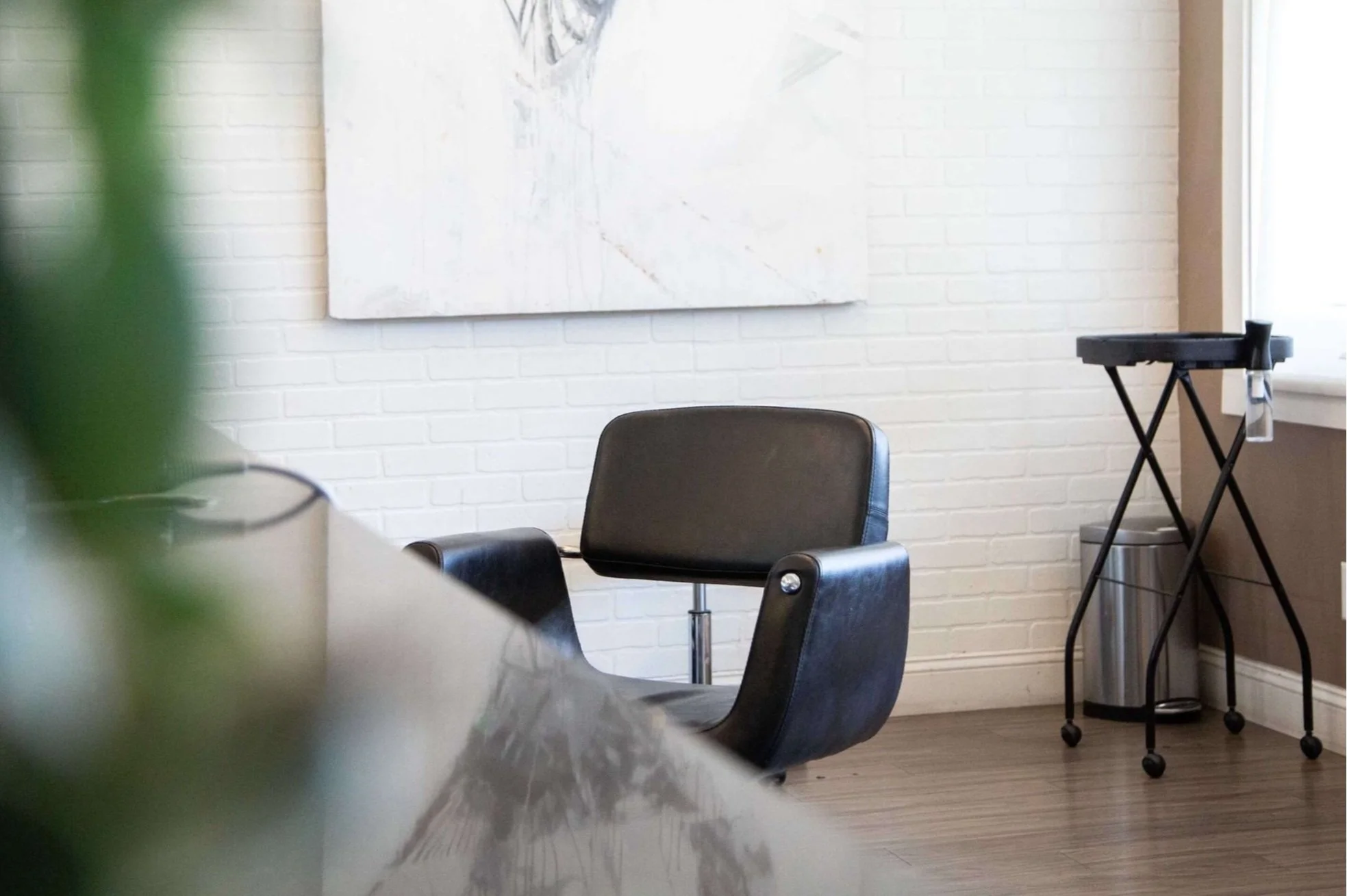 Empty black leather chair in a room with white brick wall, abstract painting, floor, and a black rolling cart near window.