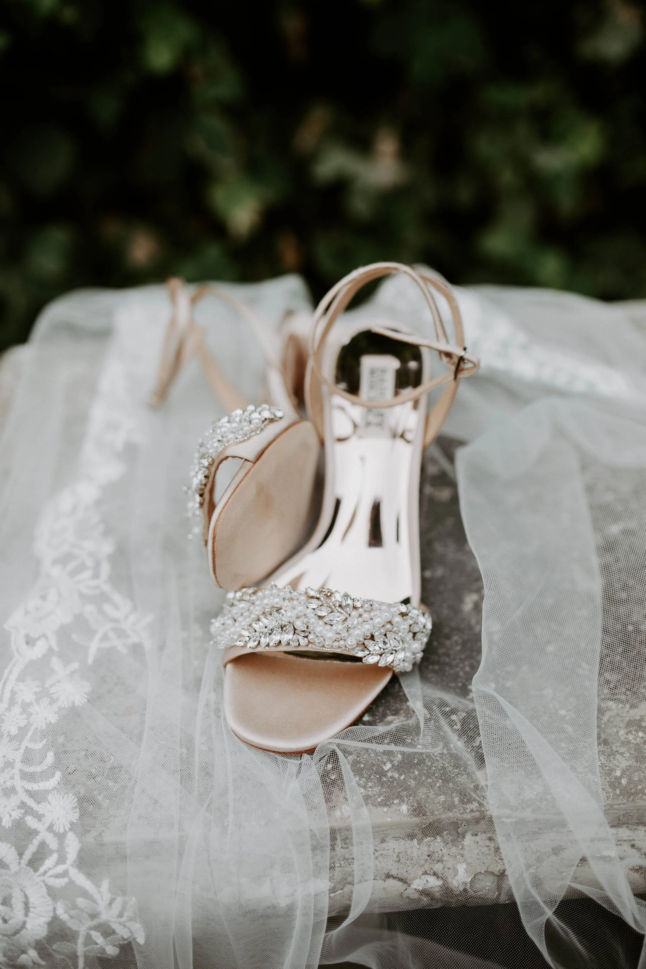 Close-up of elegant, bejeweled wedding shoes with ankle straps, resting on a lace fabric. The shoes are nude-colored with sparkling embellishments on the straps and toe area.