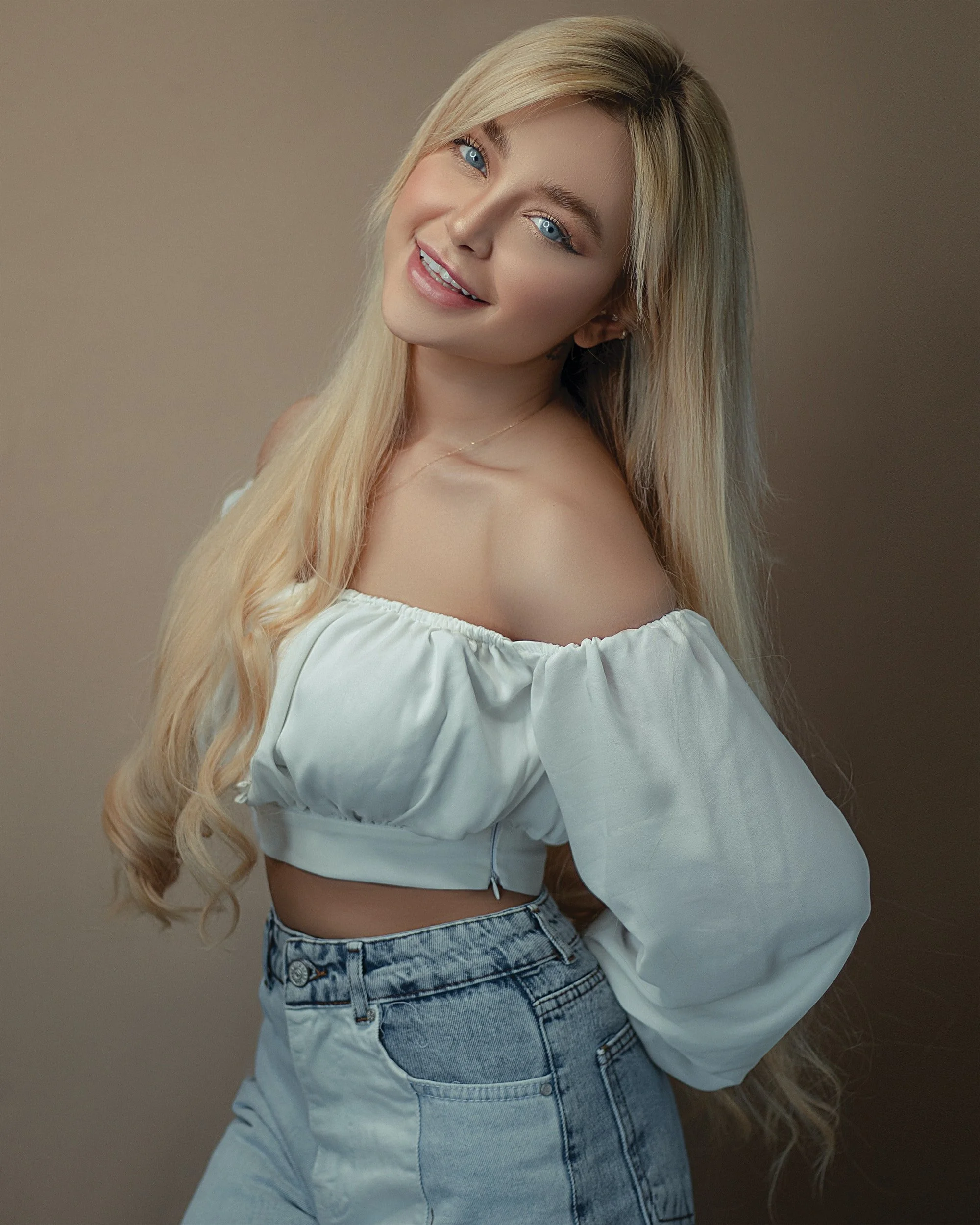 A smiling blonde woman with blue eyes wearing an off-the-shoulder white crop top and light wash jeans, posing against a plain beige background.