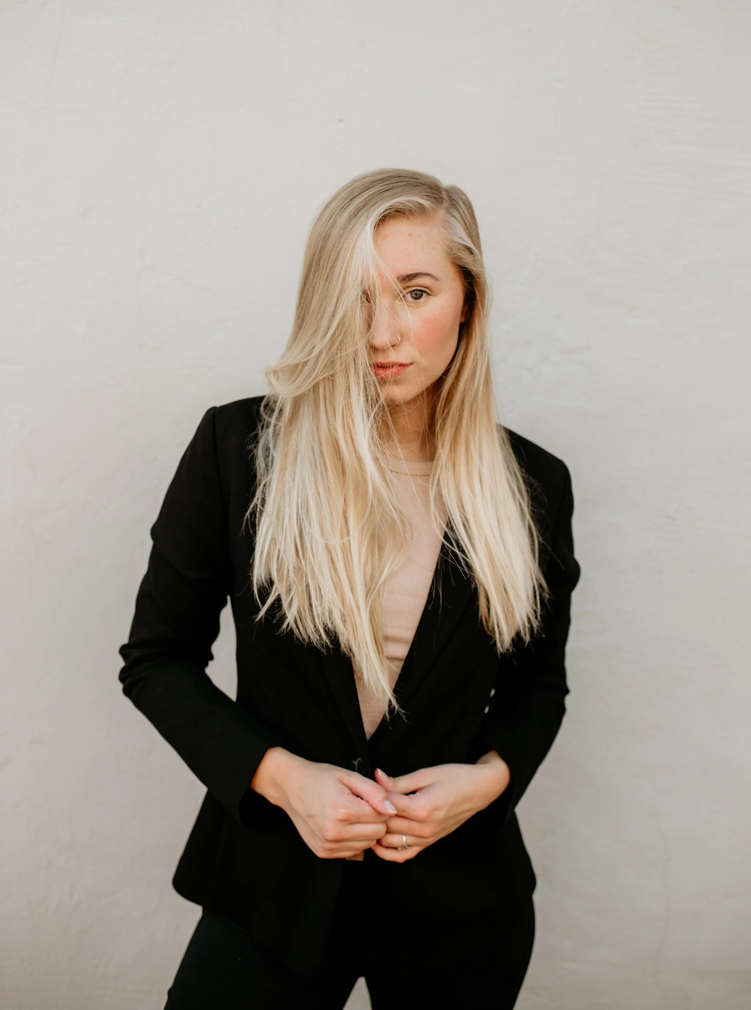 A young woman with long blonde hair, wearing a black blazer, standing against a plain light-colored wall, with a neutral expression.