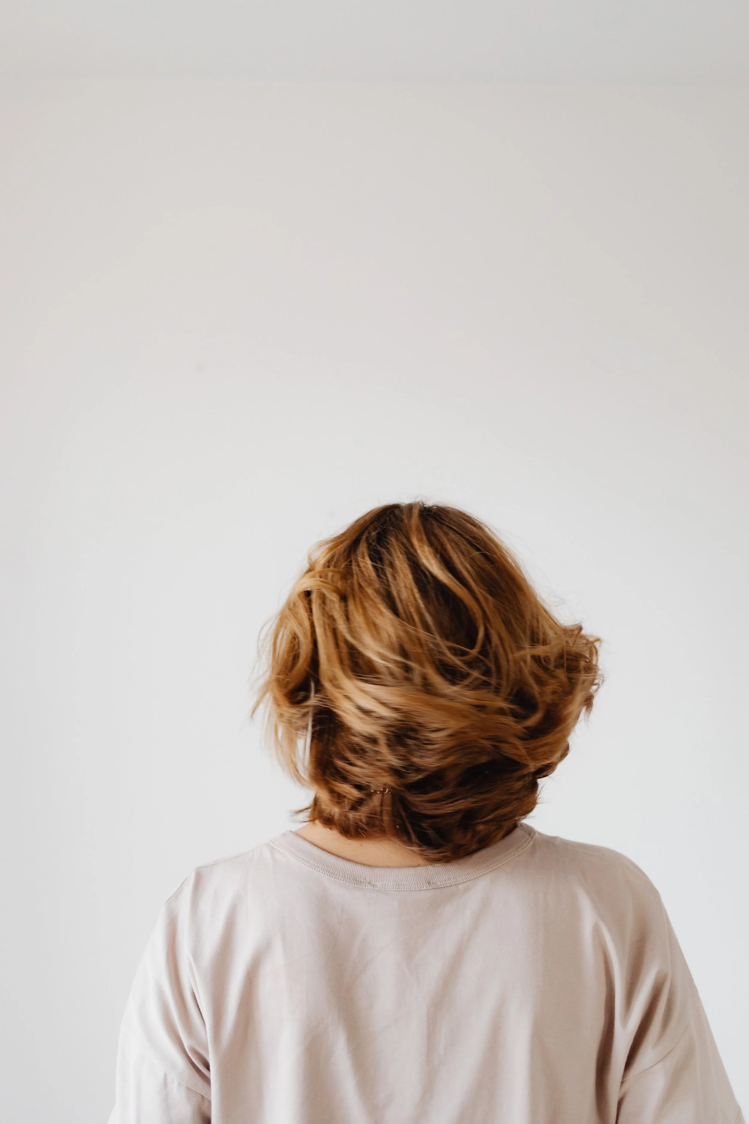 Back view of a person with shoulder-length wavy light brown hair wearing a beige shirt against a plain white wall.