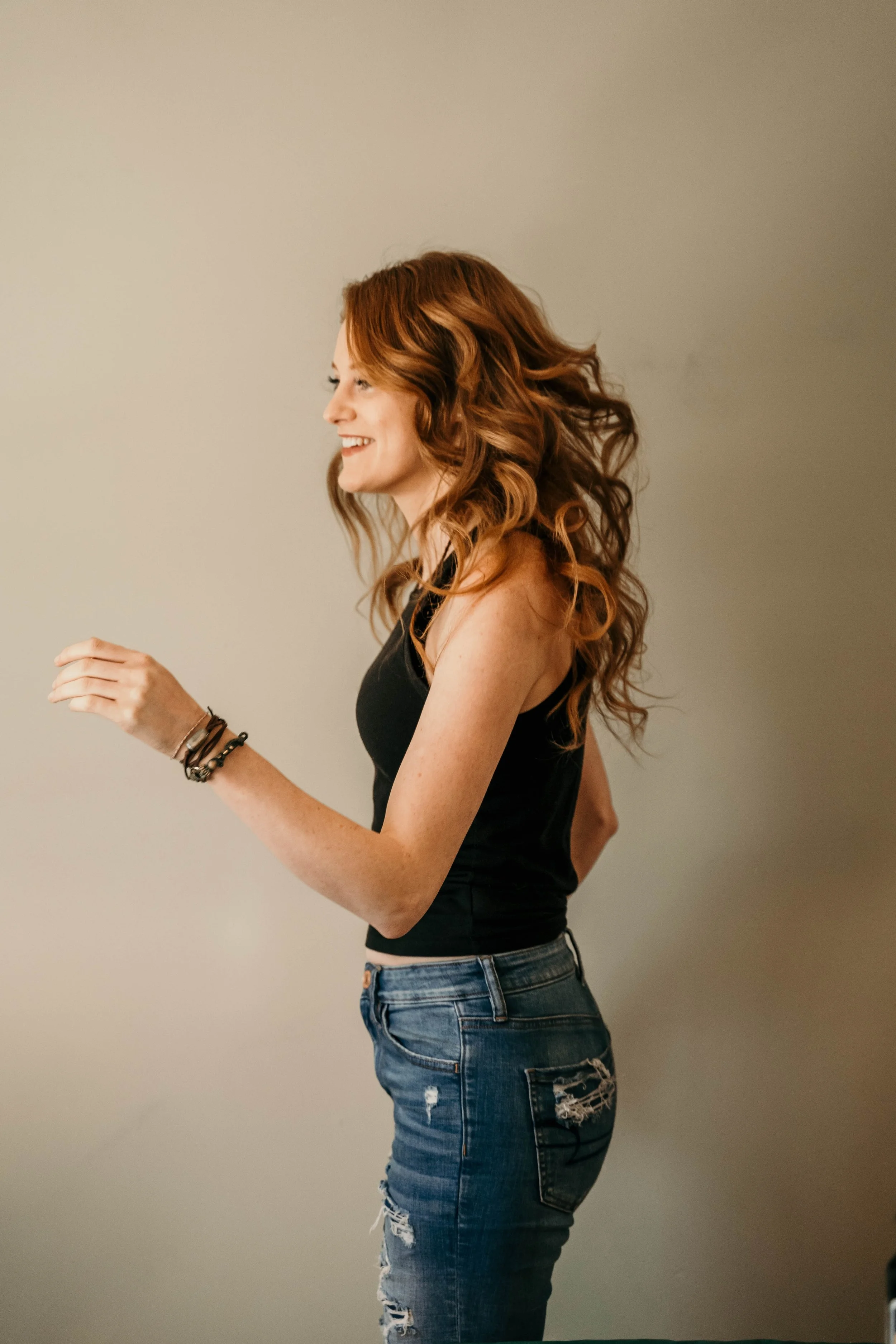 A woman with curly red hair wearing a black sleeveless top and ripped blue jeans, smiling and looking to her left against a plain light-colored background.