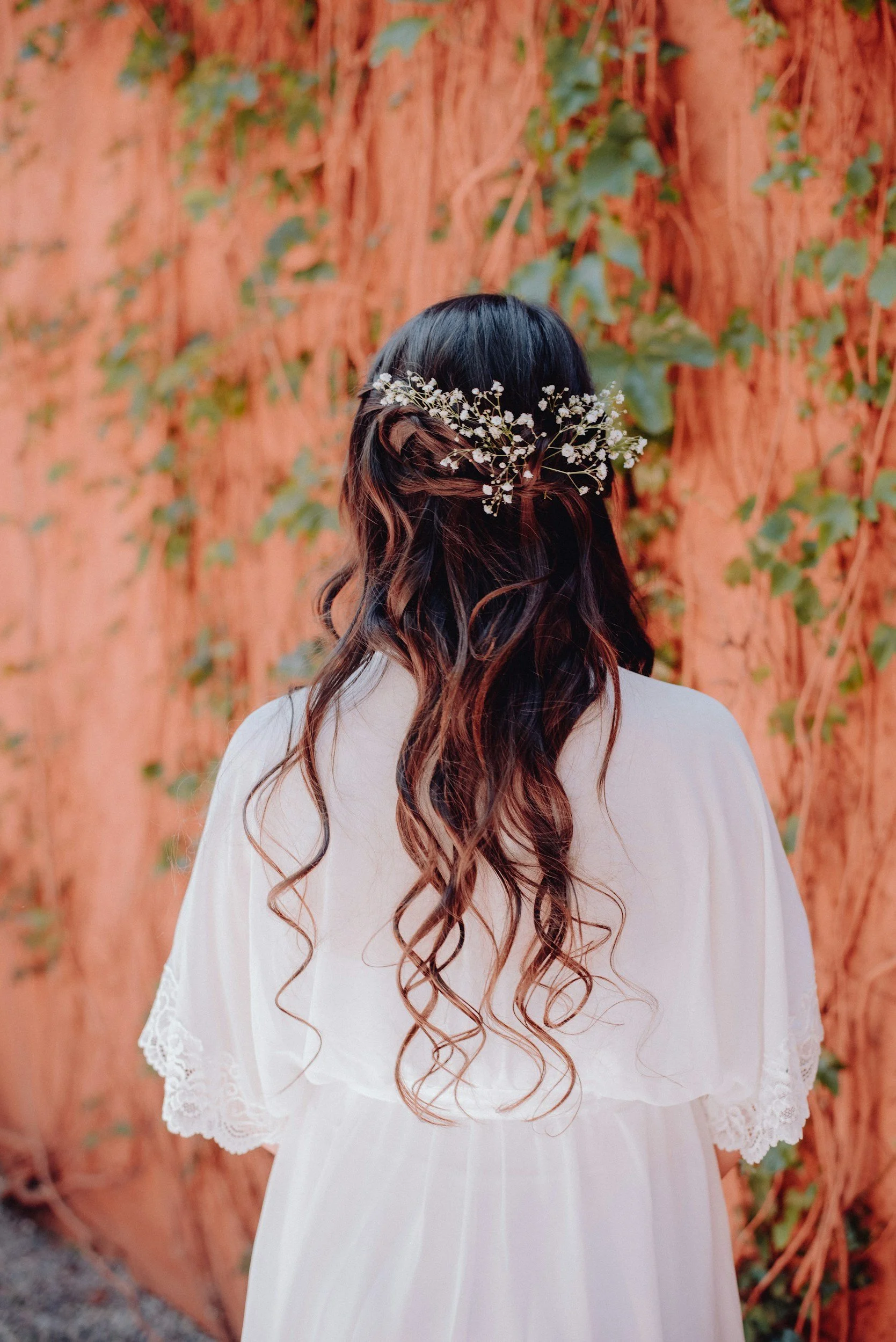 Back view of a woman with long, curly brown hair, wearing a white dress with lace details, and a floral headpiece with small white flowers, standing in front of a red, woody-textured wall covered with green ivy leaves.