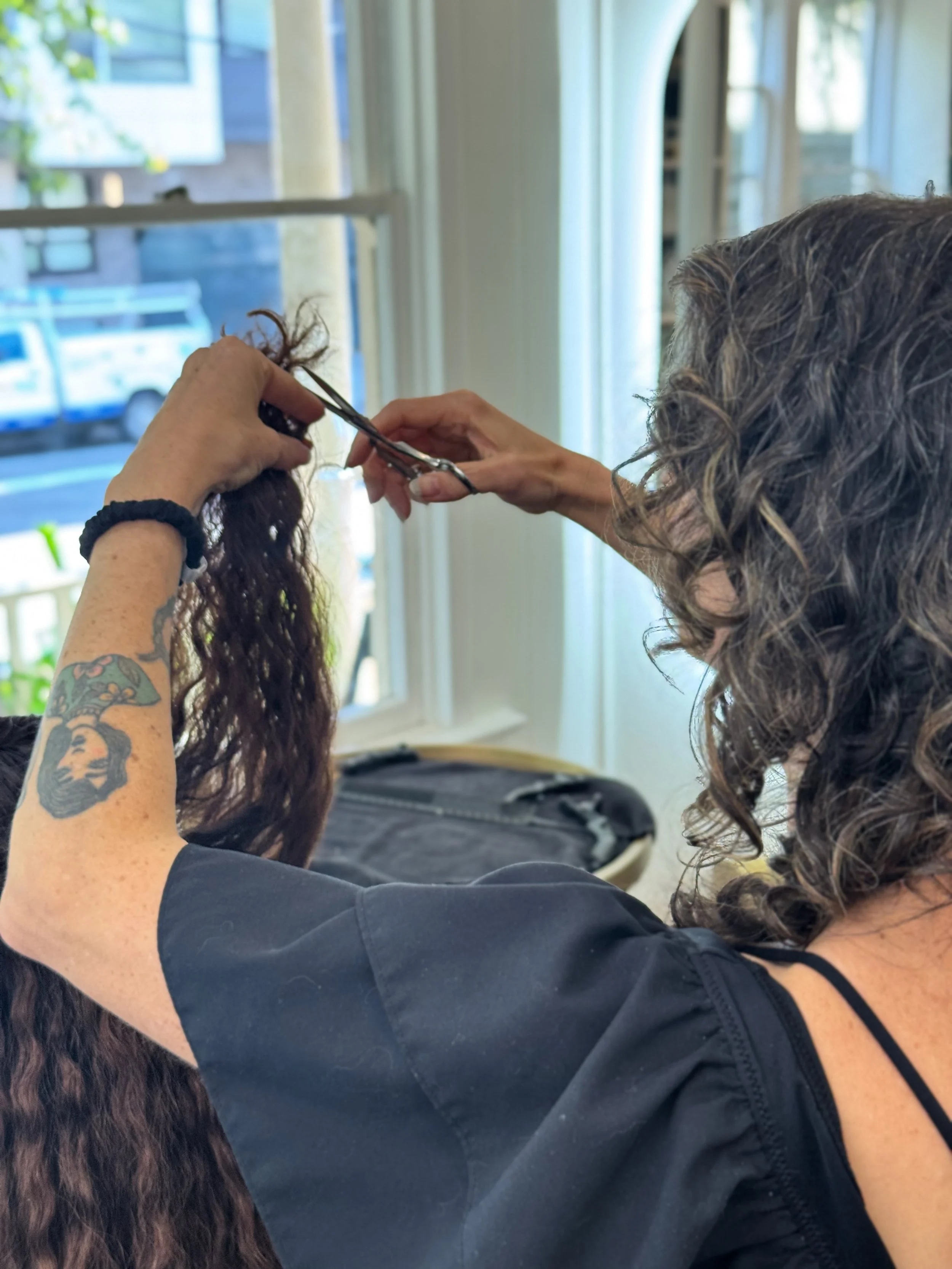 A hairstylist with curly hair and a tattoo on her arm is cutting a client's curly hair with scissors inside a bright room near a large window.