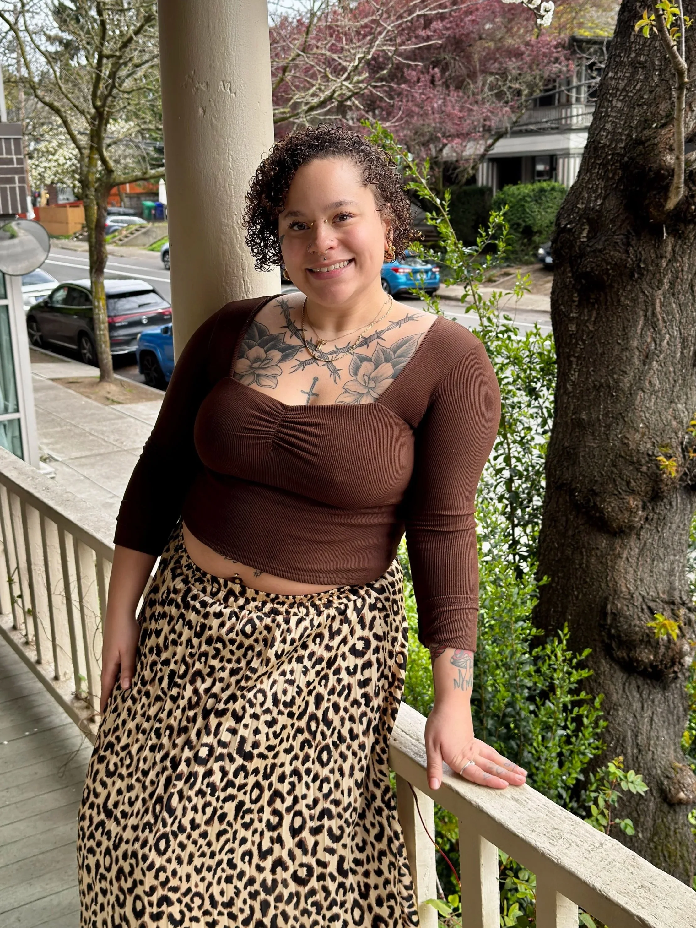 A young woman with curly hair, tattoos, and a nose piercing standing on a porch, smiling, with trees and parked cars in the background.