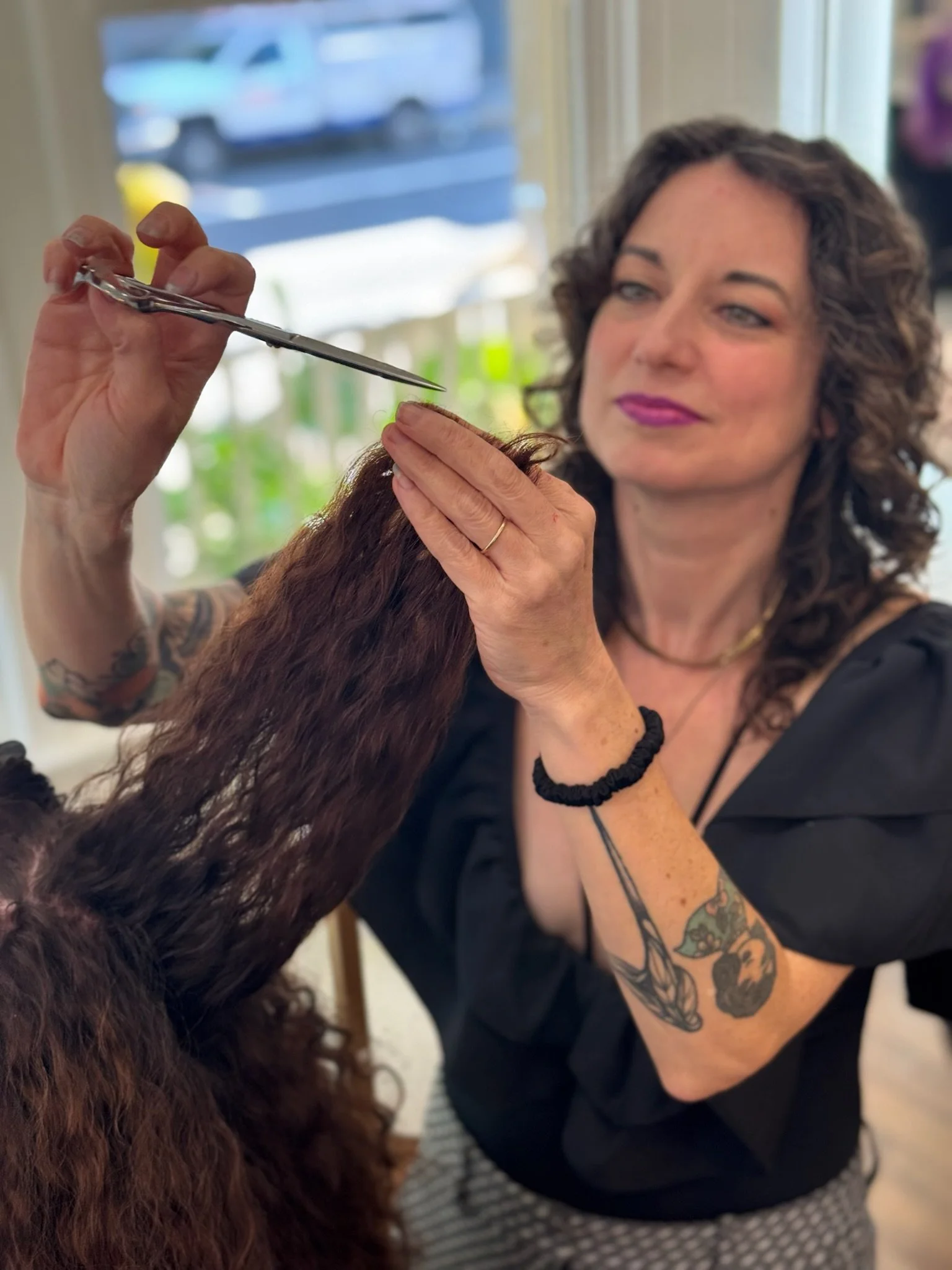 A woman with tattoos and curly hair is grooming a long, curly-haired dog with scissors indoors by a window.