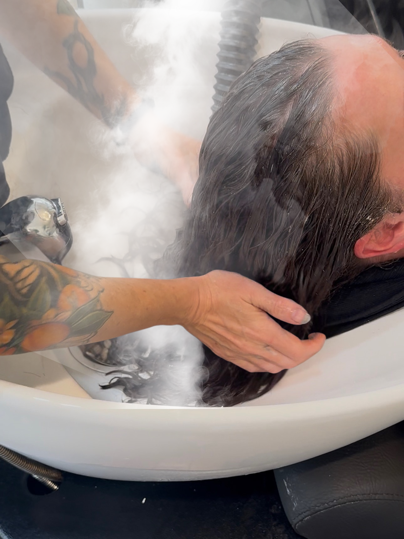 Person receiving a head shampoo at a salon or spa, with steam and shampoo foam visible on the hair and scalp.