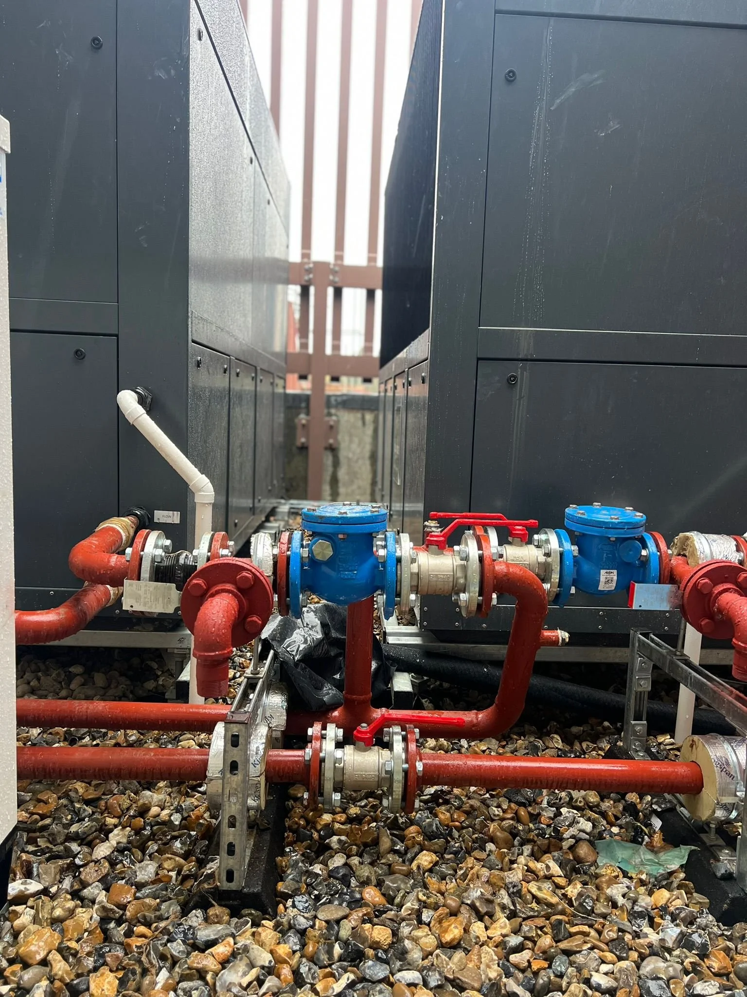 View of industrial water or HVAC piping system with red and black pipes, valves, and meters located between two large black equipment units on a gravel surface.