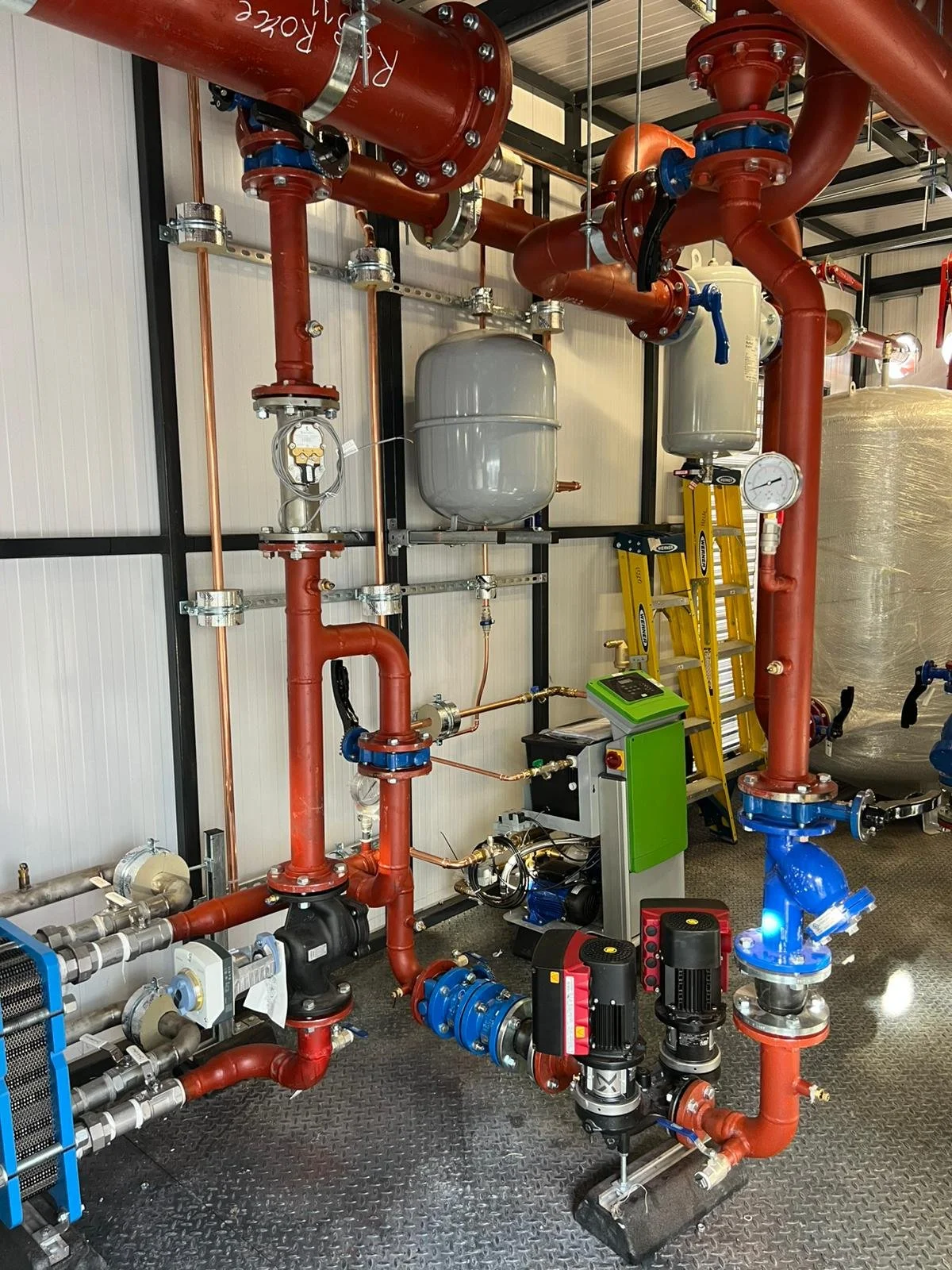 Industrial plumbing system with red pipes, pressure gauges, a gray expansion tank, a ladder, and control equipment in a utility room.