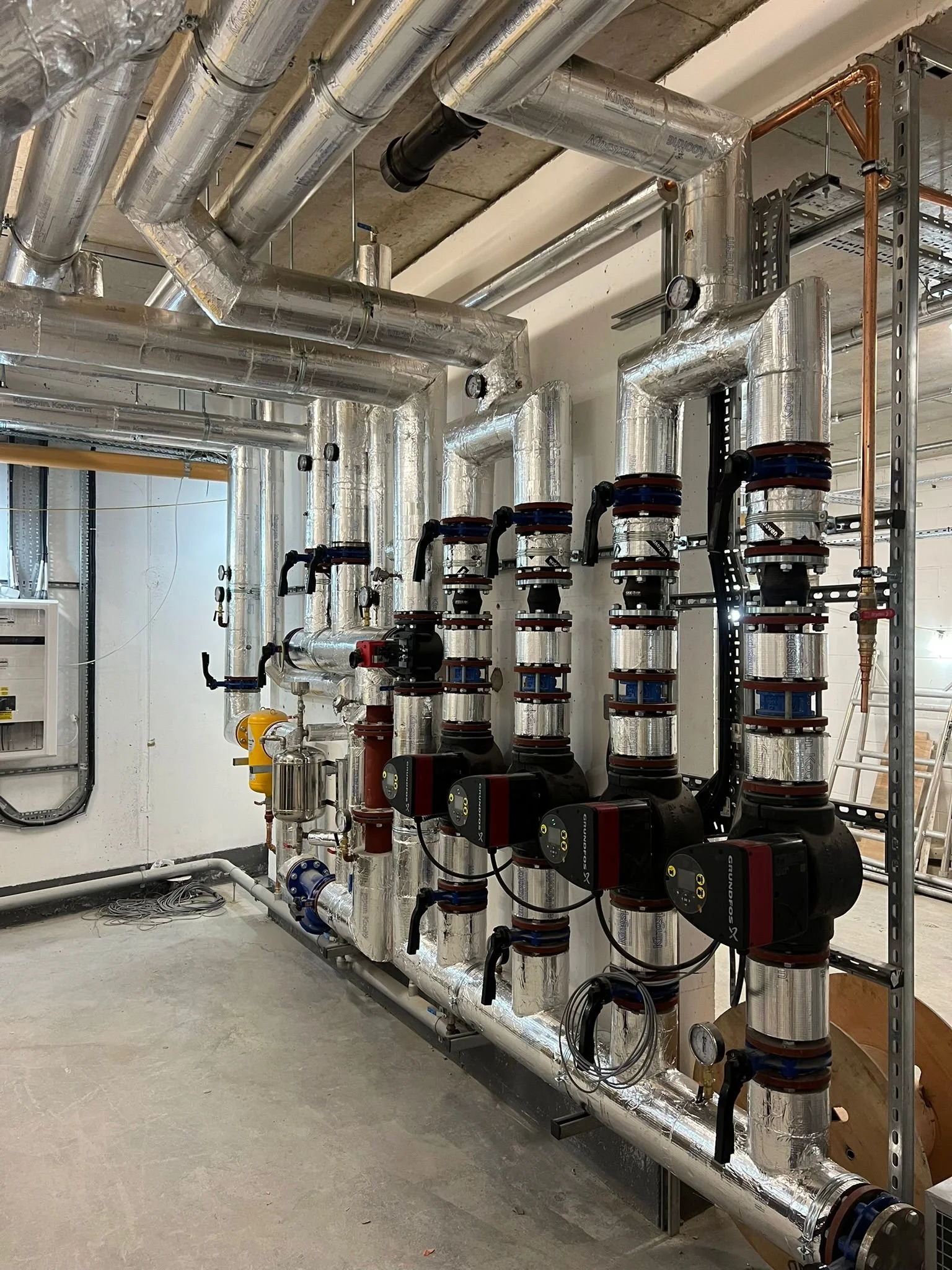 Industrial mechanical room with insulated pipes, pumps, and gauges for heating or cooling systems.