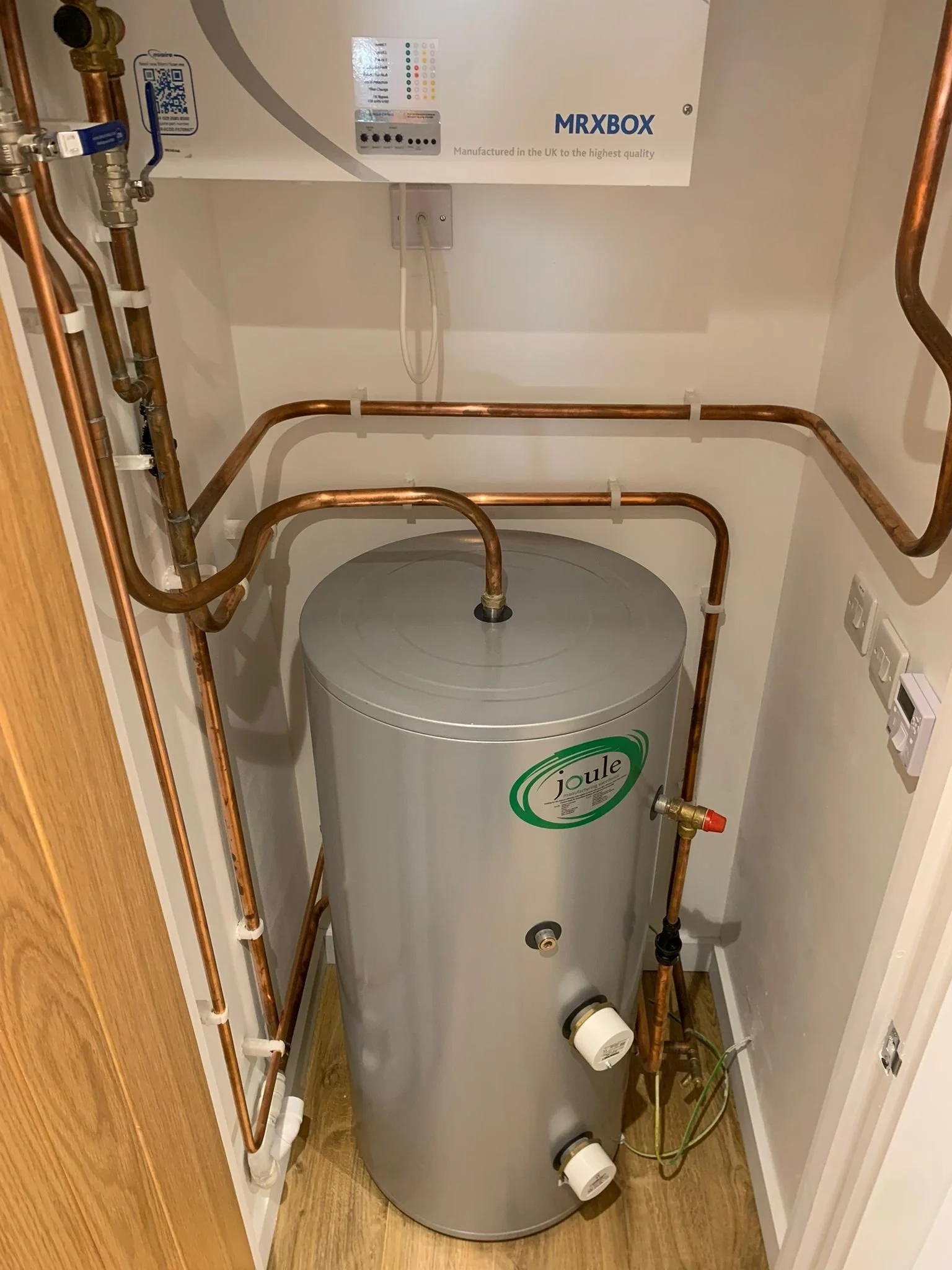 A water heater with copper pipes connected to it, mounted in a small utility closet, with control valves and labels. The closet has white walls, wooden flooring, and an electrical panel above.