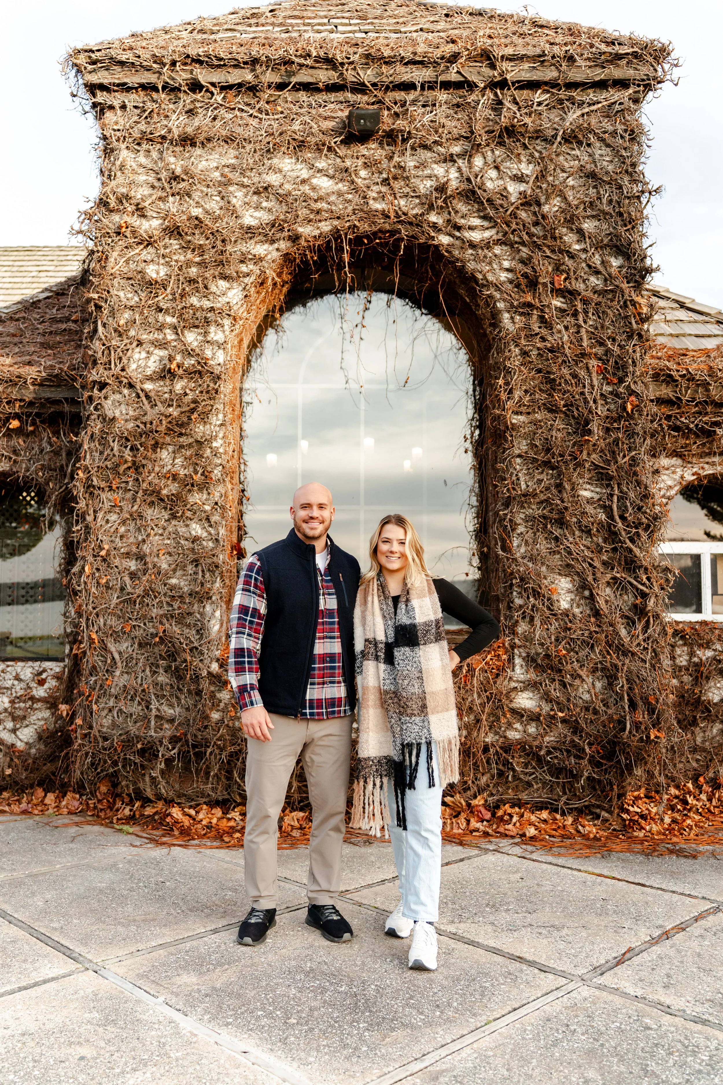 A man and woman standing in front of a large archway made of dried vines and branches, outside on a sidewalk during fall with trees and a building in the background.