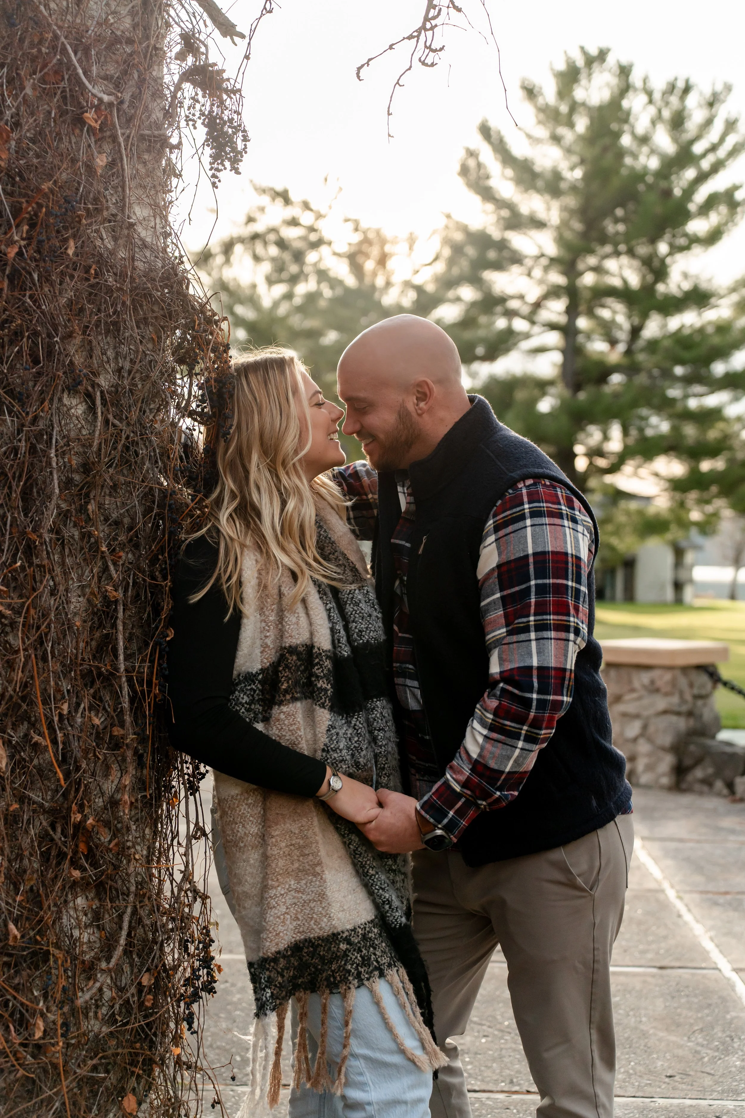 A couple standing close and holding hands, smiling, outdoors during sunset, with trees in the background.