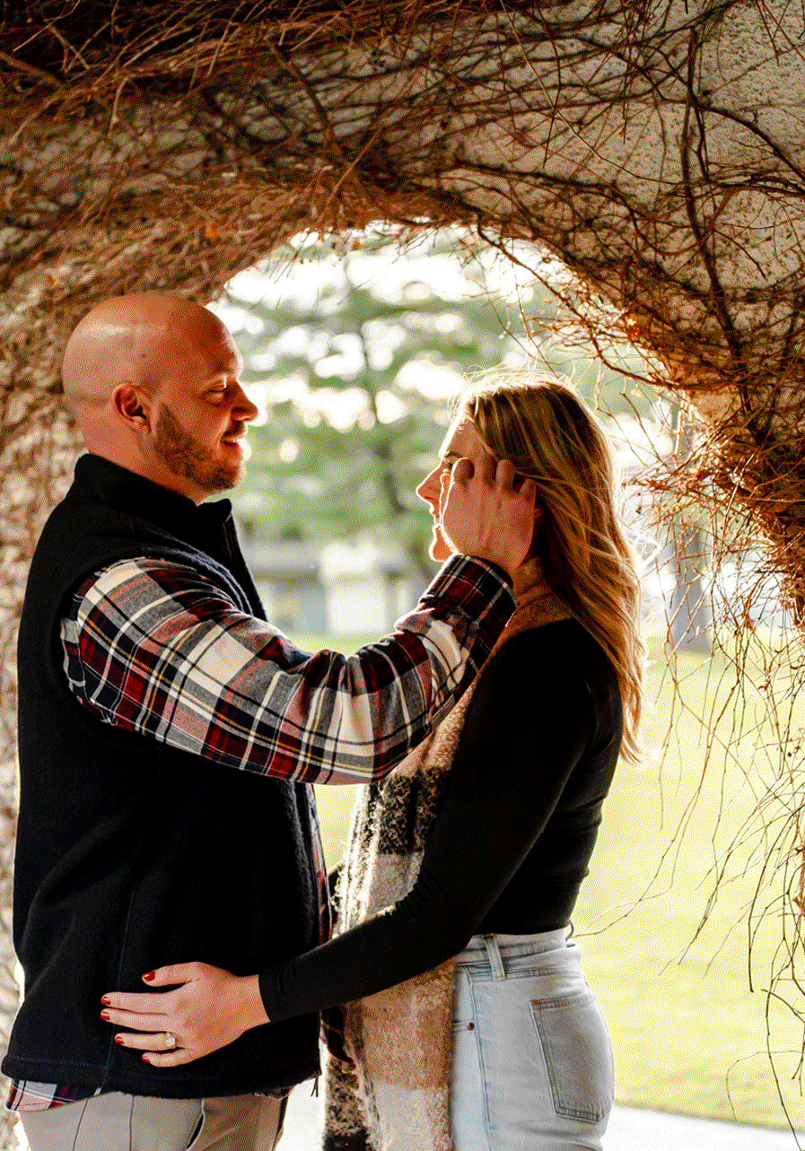 A man tenderly holds a woman's face and smiles at her, standing beneath a large tree with bare branches. The woman is embracing the man with one hand on his waist, and they look into each other's eyes during sunset.