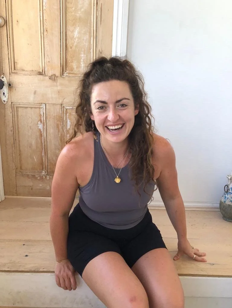A woman with curly brown hair smiling and sitting on a wooden step inside a house, wearing a sleeveless gray top and black shorts.