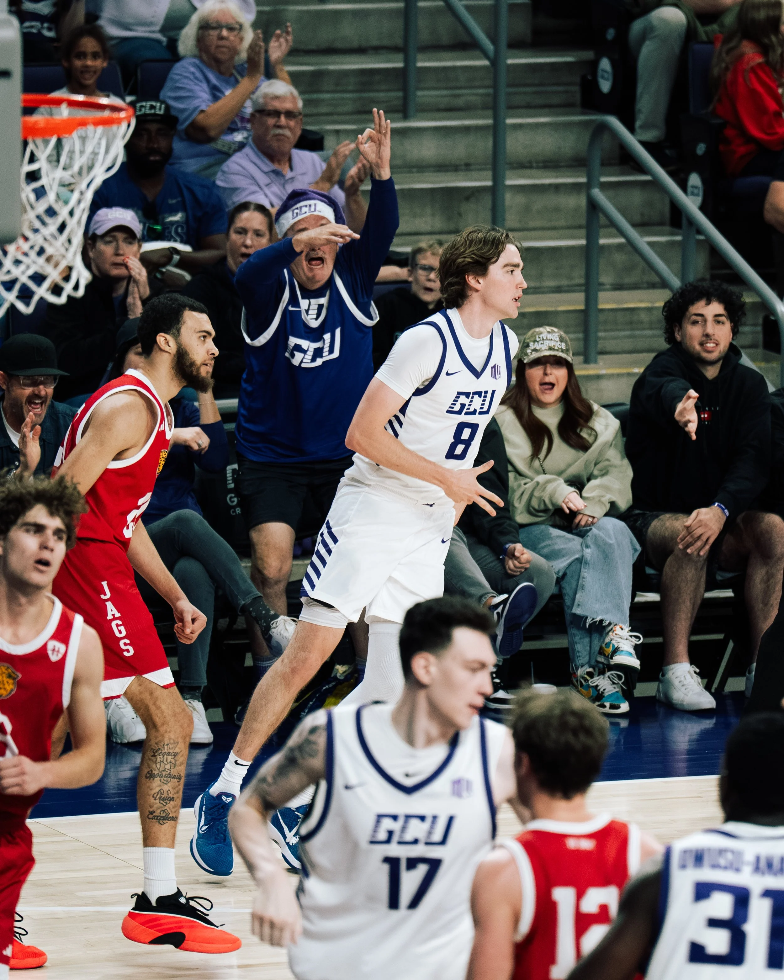 A basketball game with players from GCU and JAGS teams, playing on the court with spectators in the background. One player in a white jersey is running, while a fan in GCU gear is showing excitement near the bleachers.