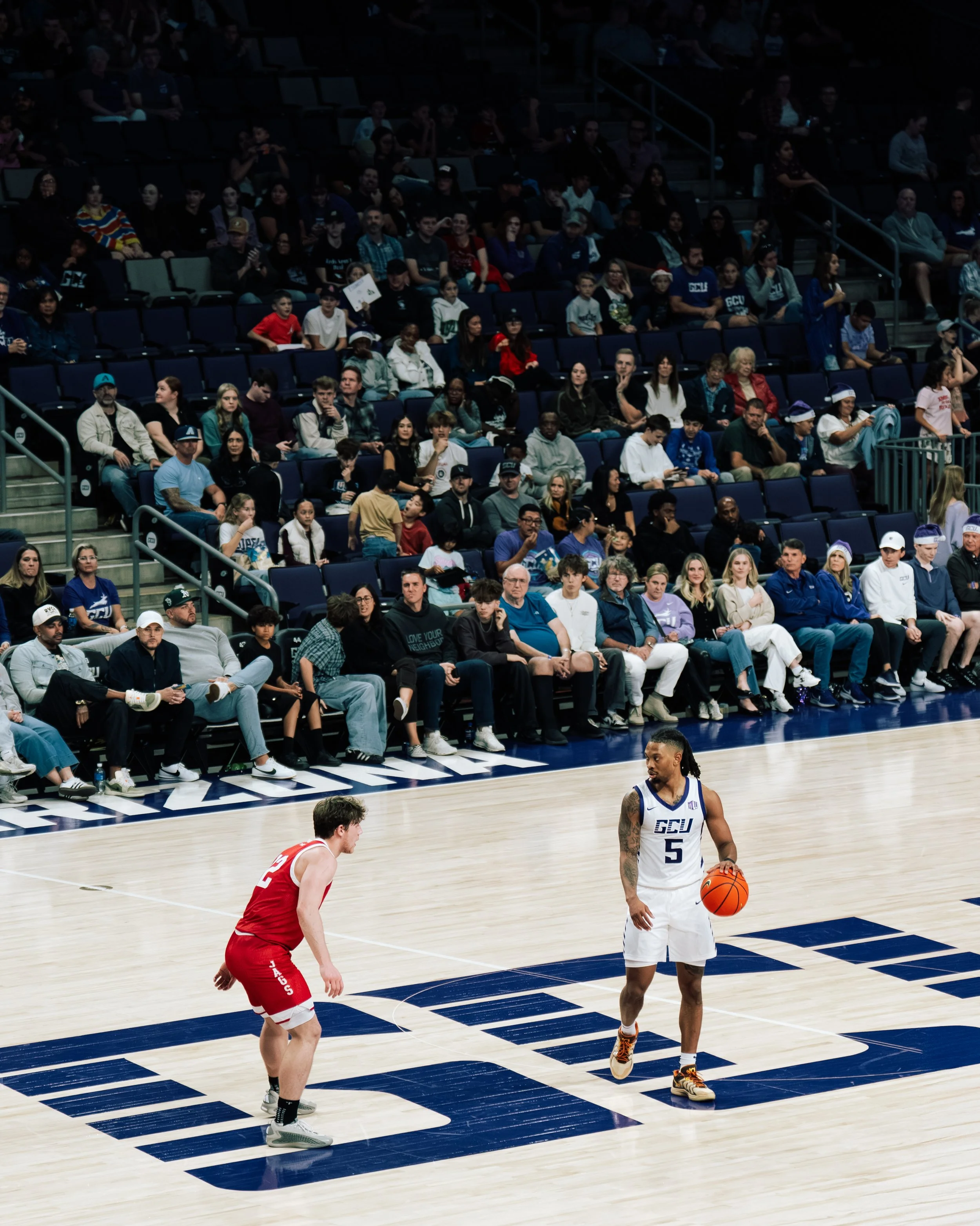 A basketball game with one player in a red jersey and another in a white jersey with the number five, on a gymnasium court with a crowd of spectators watching.