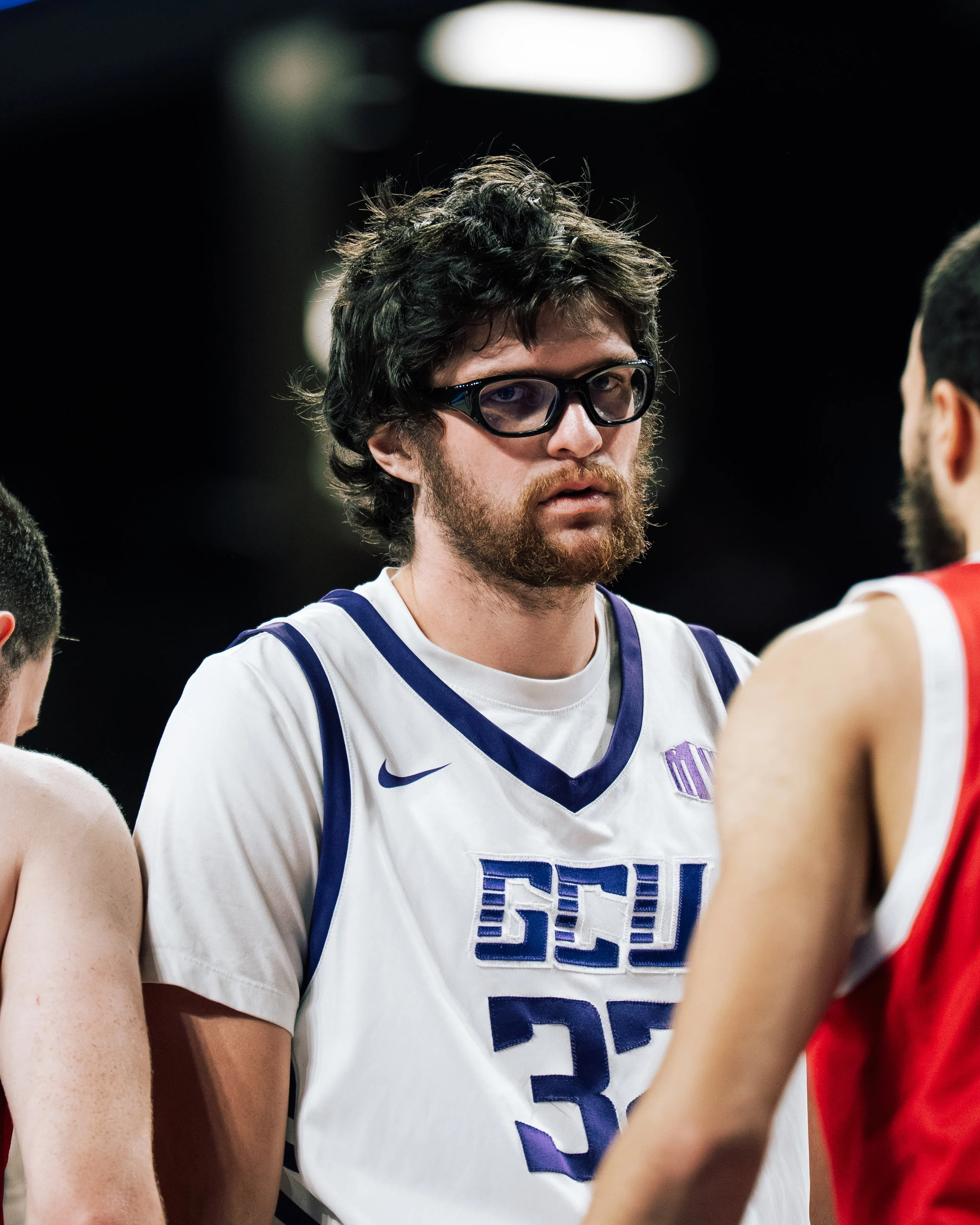 Basketball player with glasses wearing a white jersey with blue trim, standing with other players during a game.