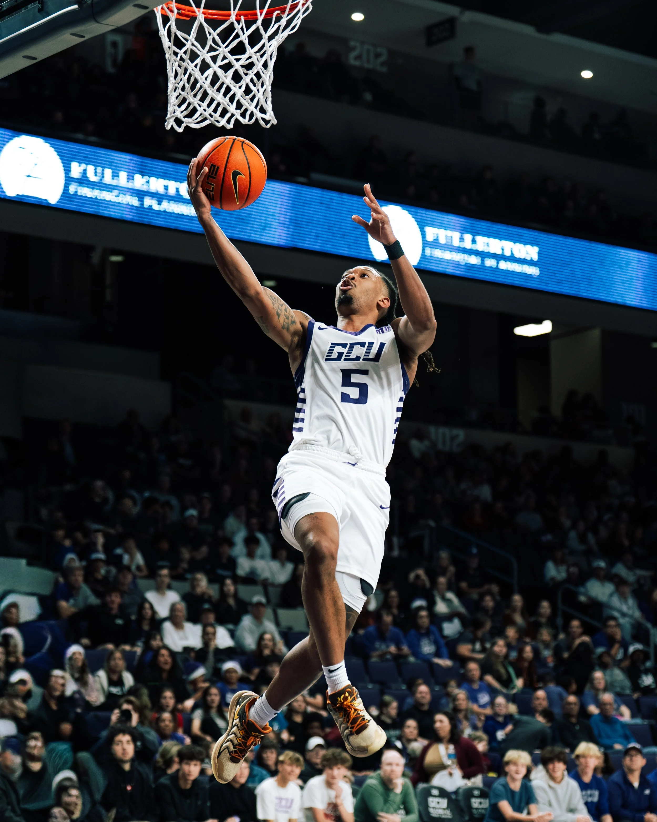 A basketball player in a white jersey with the number 5, jumping towards the hoop for a layup shot during a game with spectators in the background.