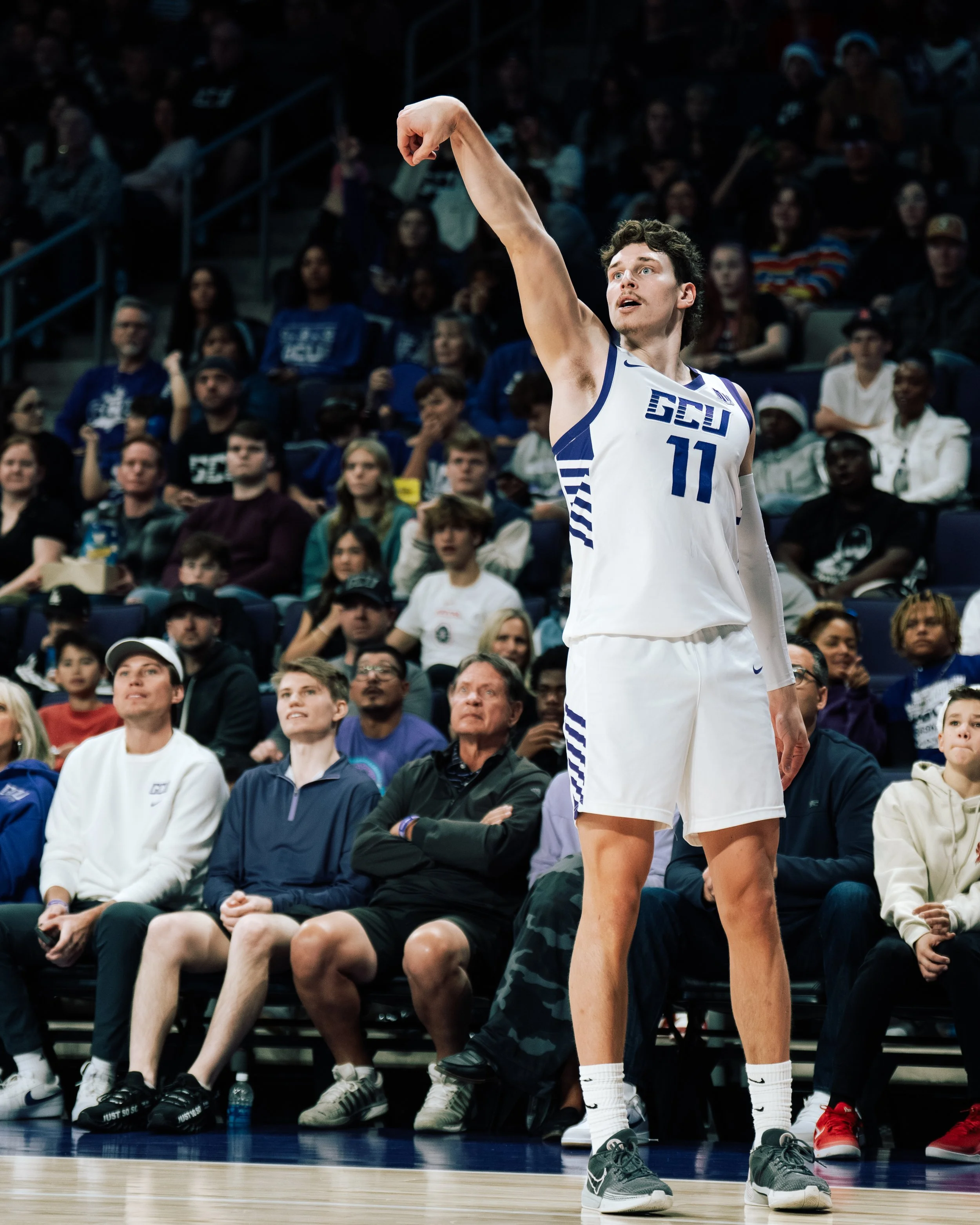 A basketball player wearing a white and blue uniform with the number 11 and the letters 'SCU' on it, taking a shot during a game. Spectators sit in the background watching.