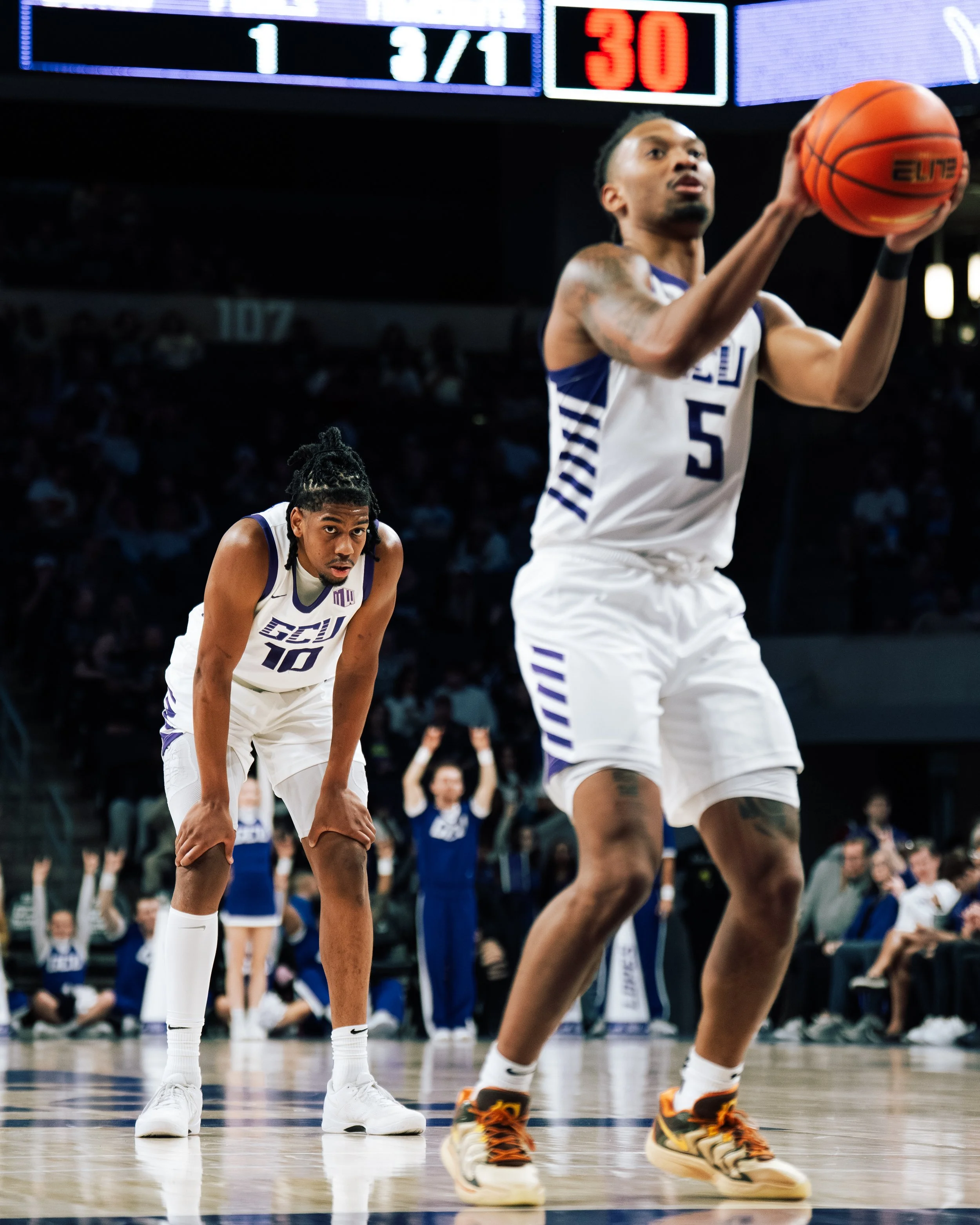 Two basketball players in white uniforms with purple accents on the court, one preparing to shoot and the other in a defensive stance, with a scoreboard overhead showing 1 and 30 seconds.
