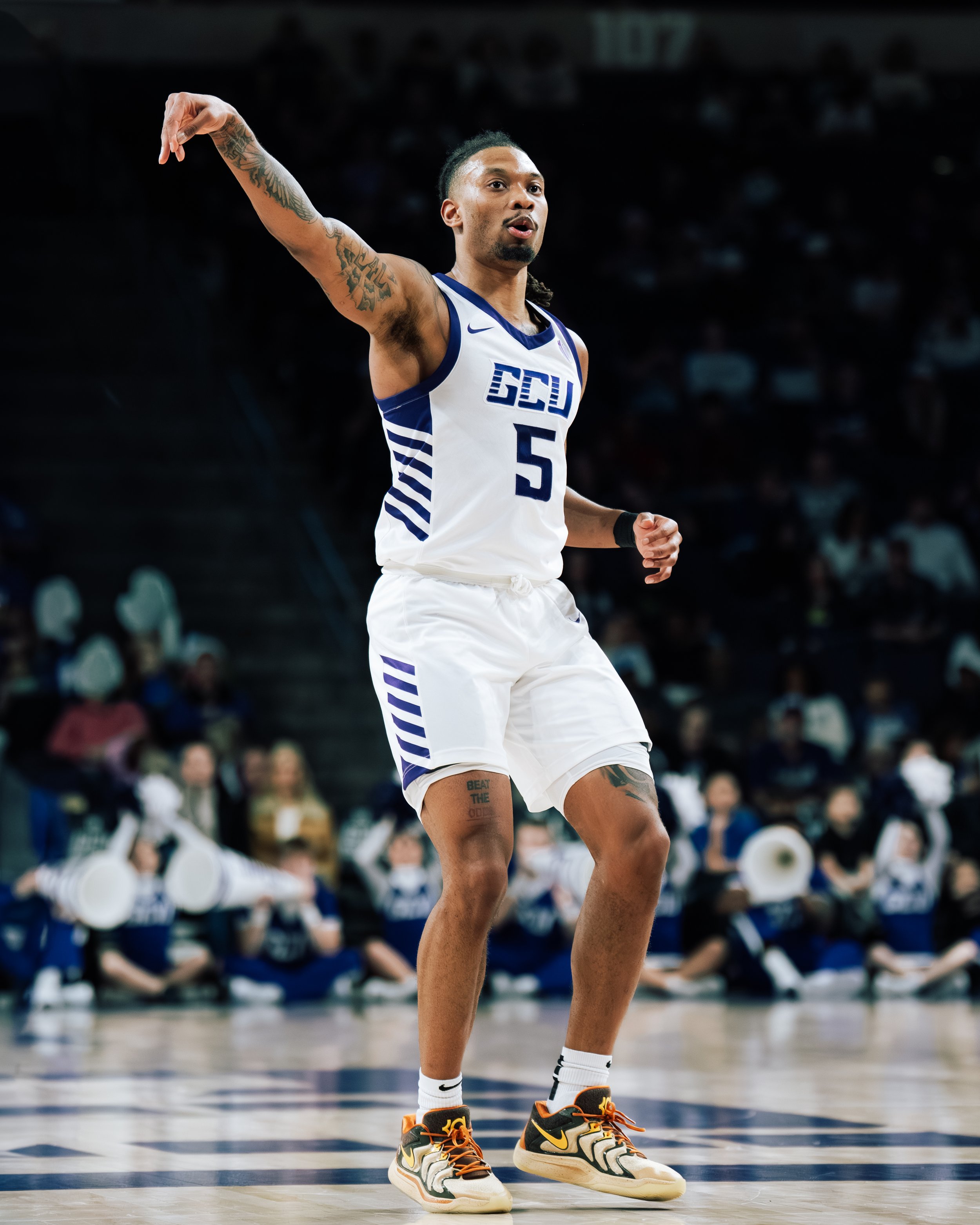 A basketball player on the court wearing a white and blue jersey with the number 5 and gang tattoos, standing with his right arm raised, during a game at a packed arena.