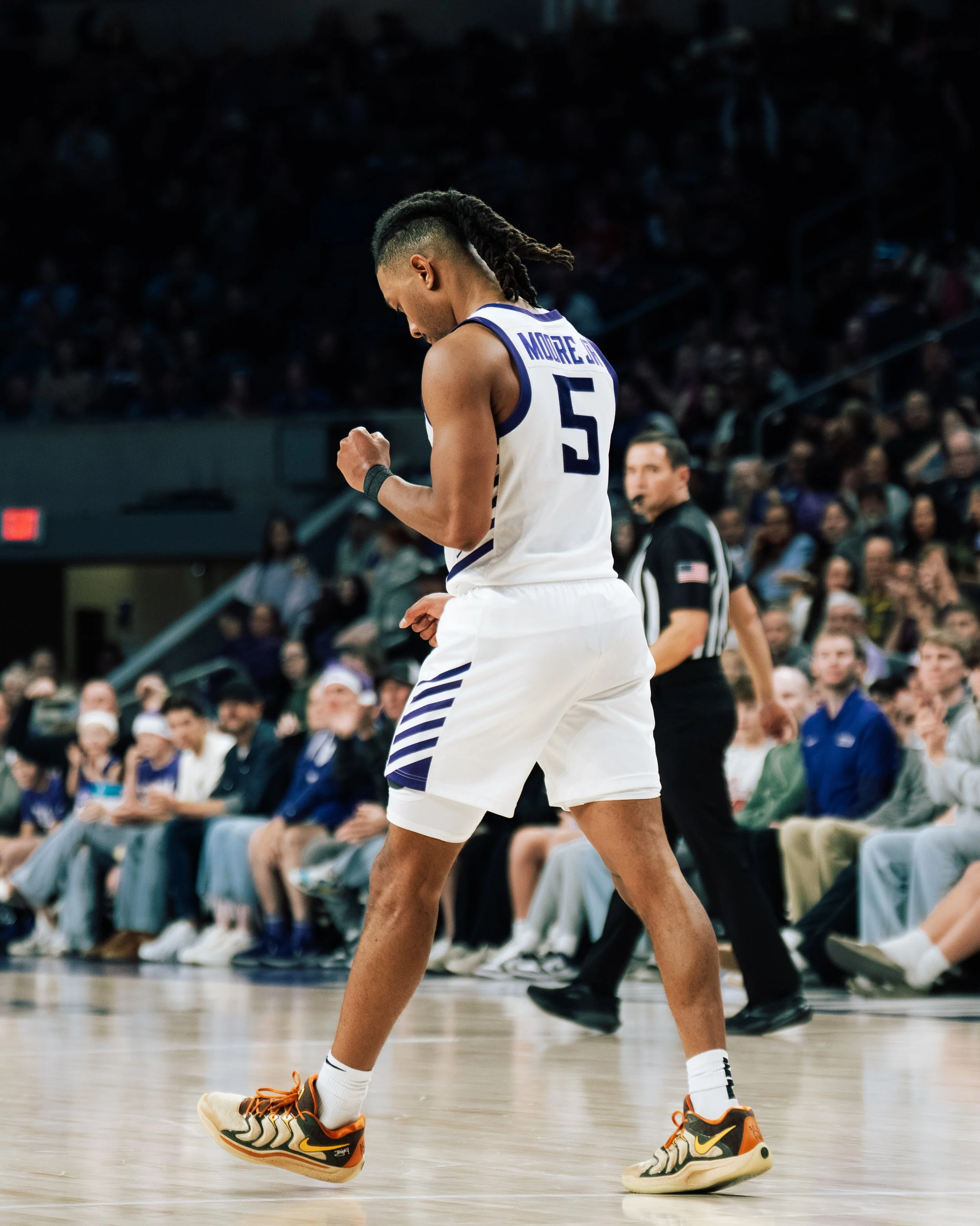 A basketball player wearing a white and blue uniform with the number 5, standing on a basketball court with a crowd in the background, looking down with clenched fists.