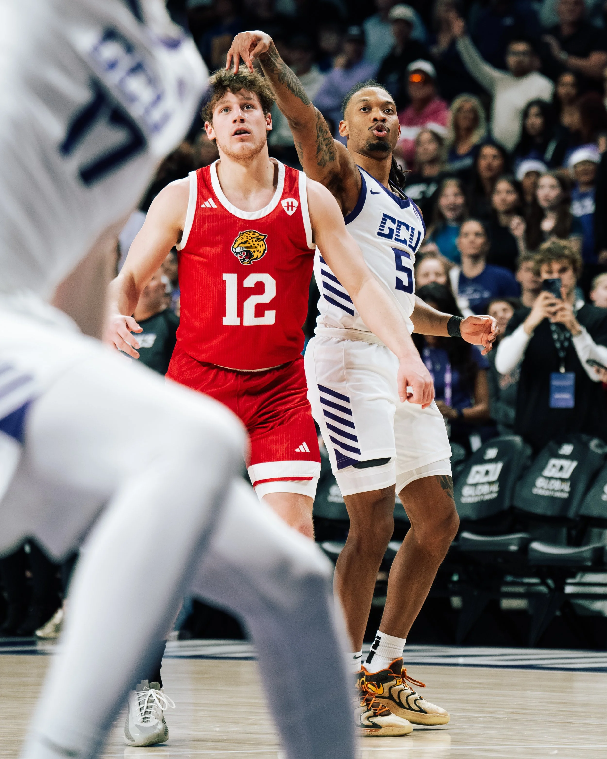 Two basketball players, one in a red jersey with number 12 and the other in a white jersey with number 5, are playing on the court during a game. The player in red is focused with his mouth slightly open, while the player in white is looking in the s
