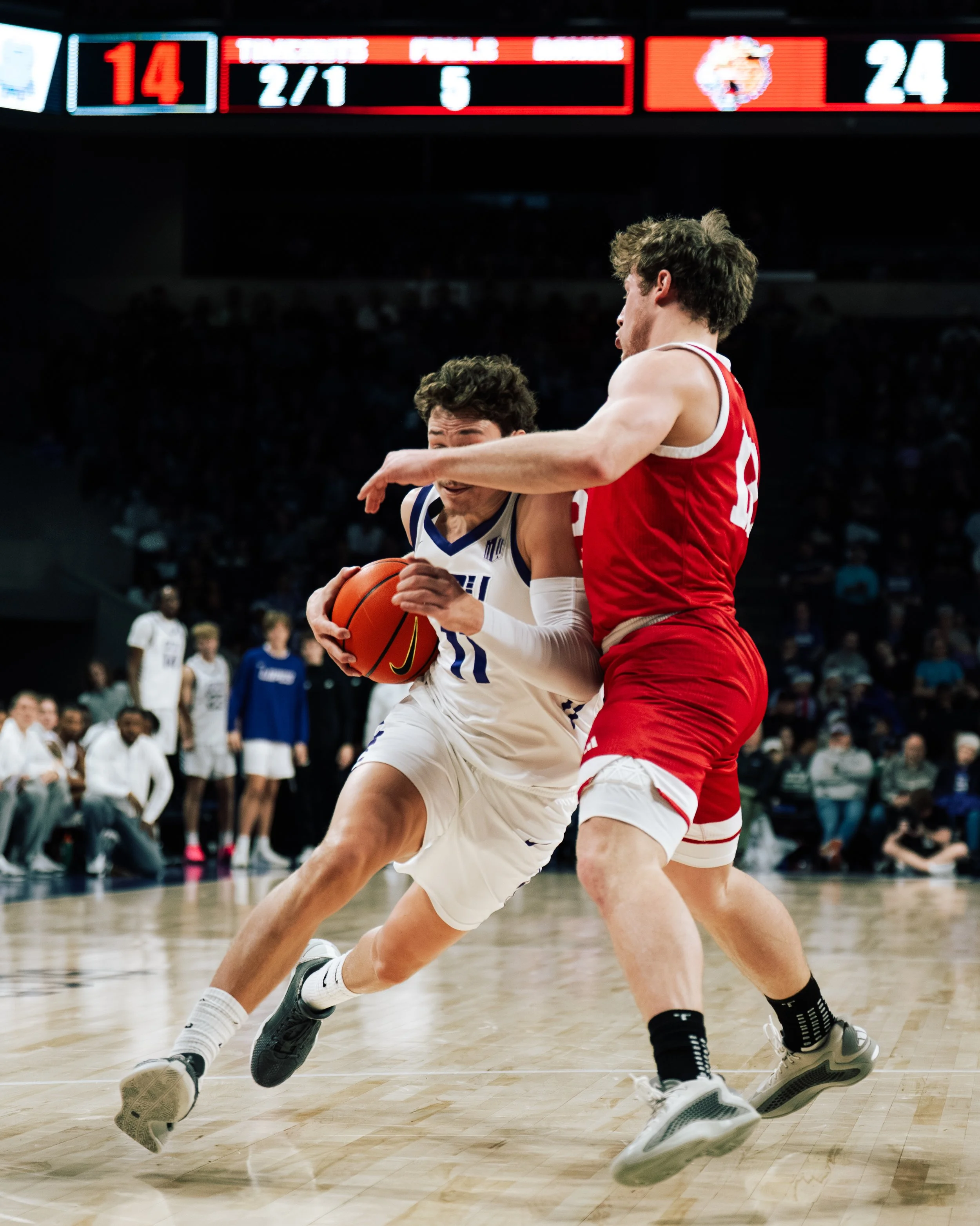 Two basketball players compete during a game, with one player in a white jersey holding the ball and a player in a red jersey defending.