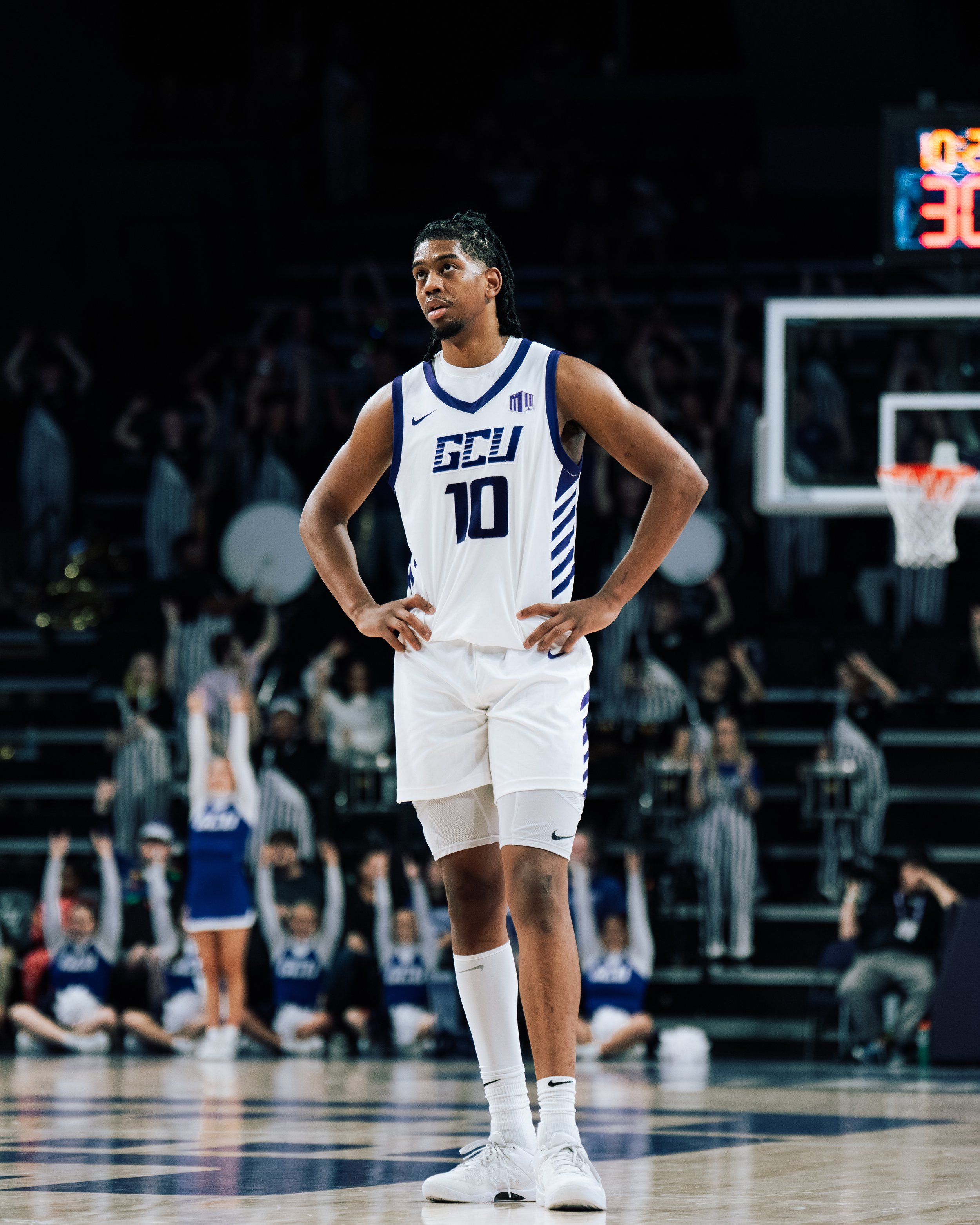 A basketball player stands on the court with hands on hips, wearing a white jersey with blue accents and the number 10, in a stadium with cheering audience and cheerleaders in the background.