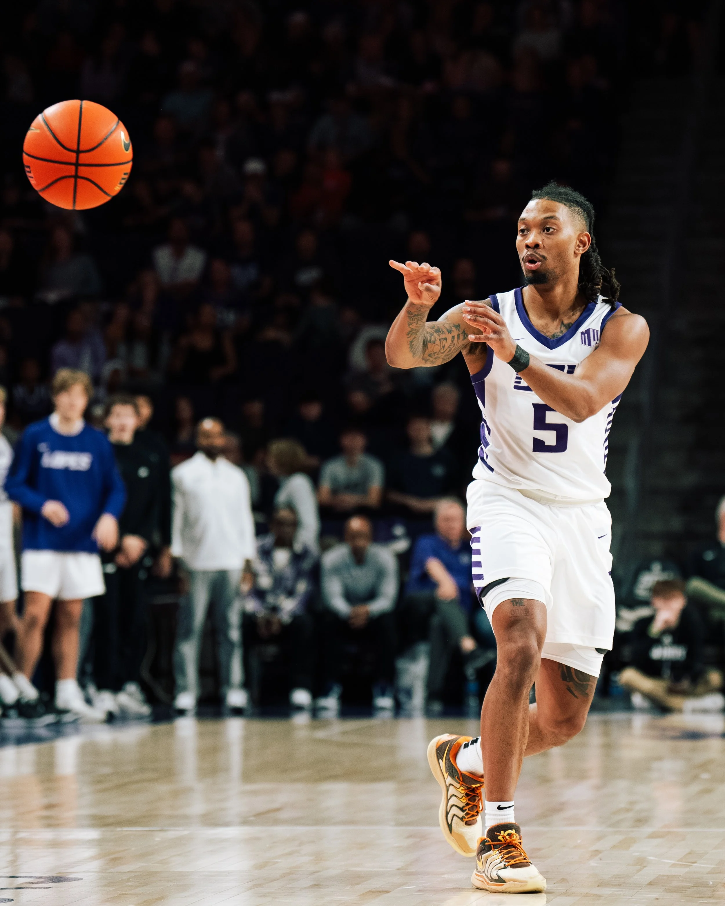 A basketball player wearing a white jersey with number 5 performs a pass on the court while a basketball is in mid-air, with an audience and other players in the background.