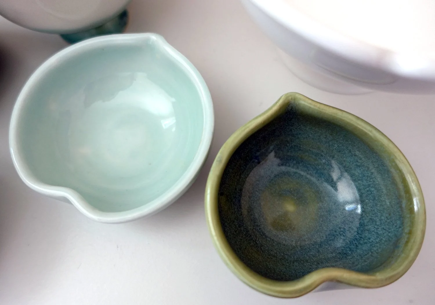 Two small ceramic bowls with uneven pouring spouts, one in mint green and the other in blue-glazed finish, placed on a white surface.