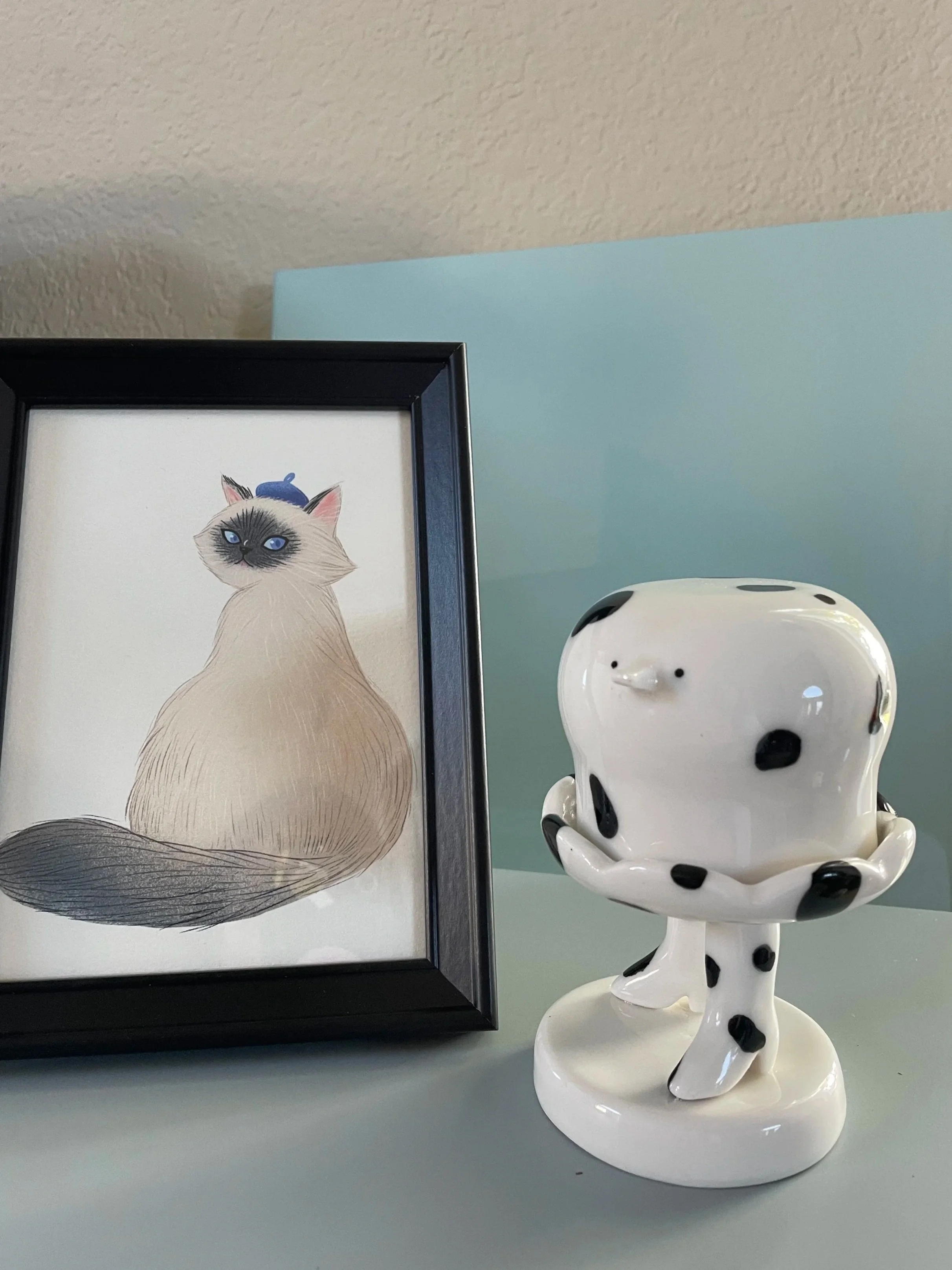 A framed illustration of a Siamese cat with blue eyes and a tiny blue hat, placed next to a ceramic sculpture of a Dalmatian with black spots and a rounded body