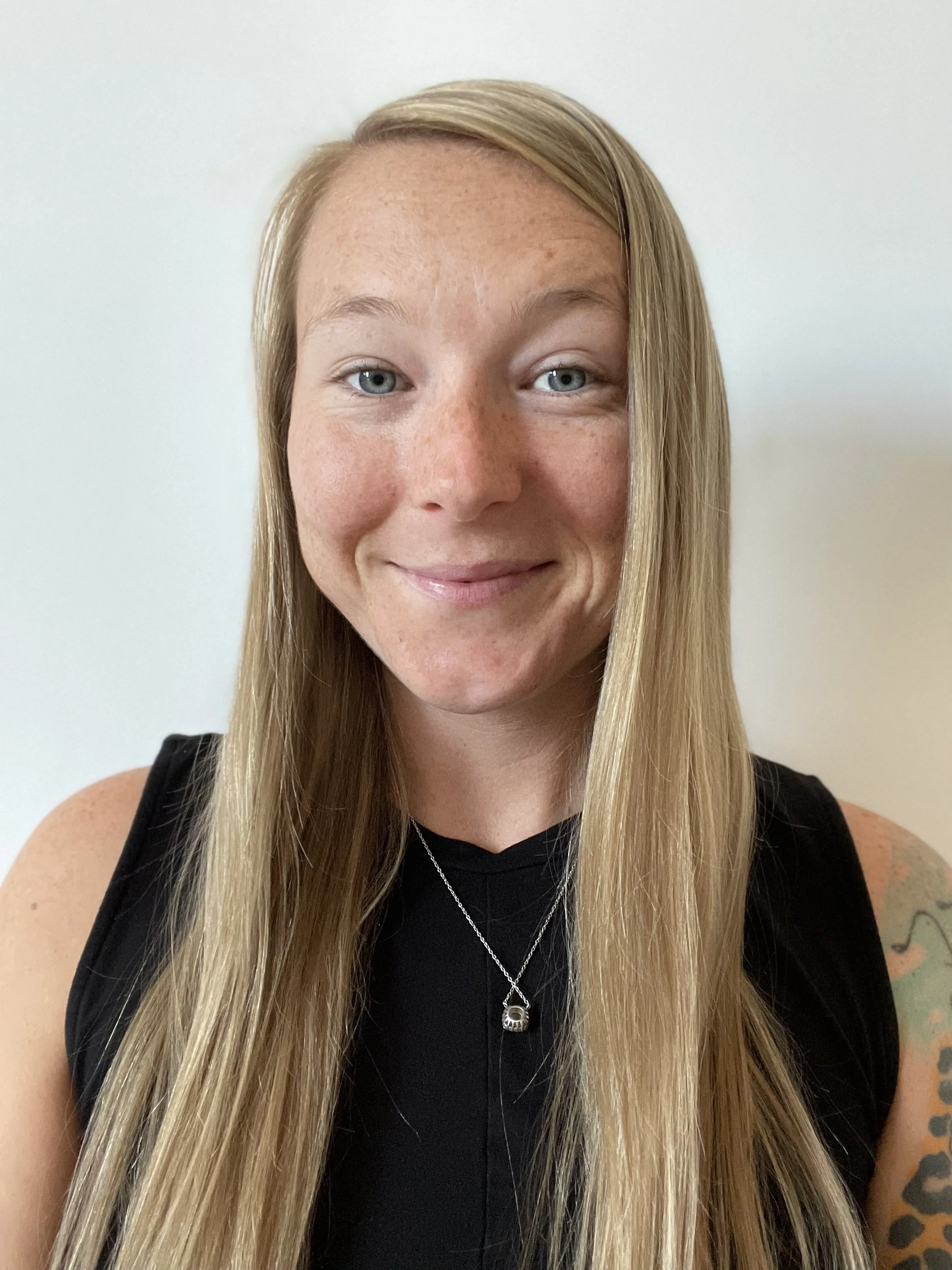 Close-up photo of a young woman with long blonde hair, blue eyes, freckles, wearing a black top, a silver skull necklace, and a tattooed arm, smiling slightly against a plain white background.
