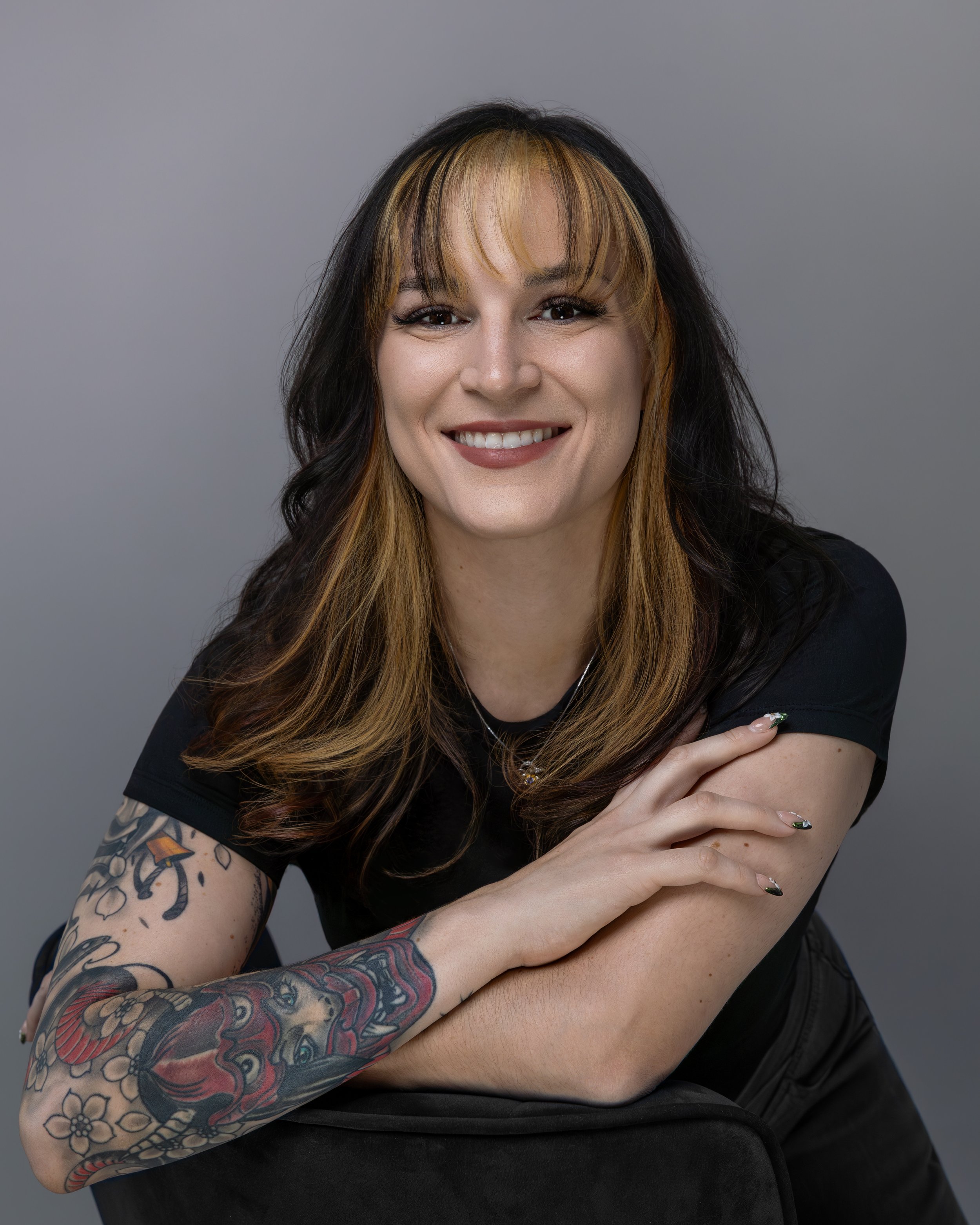 A smiling woman with shoulder-length hair, wearing a black shirt, showing tattoos on her left arm and a ring on her finger against a gray background.