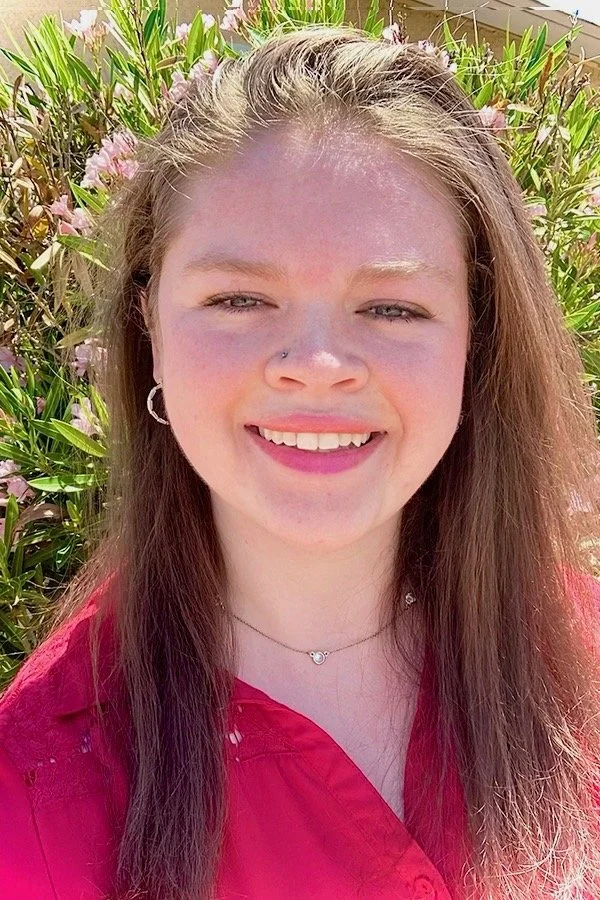 A young woman with long brown hair smiling in front of pink flowers and green foliage, wearing a red shirt and a silver necklace.