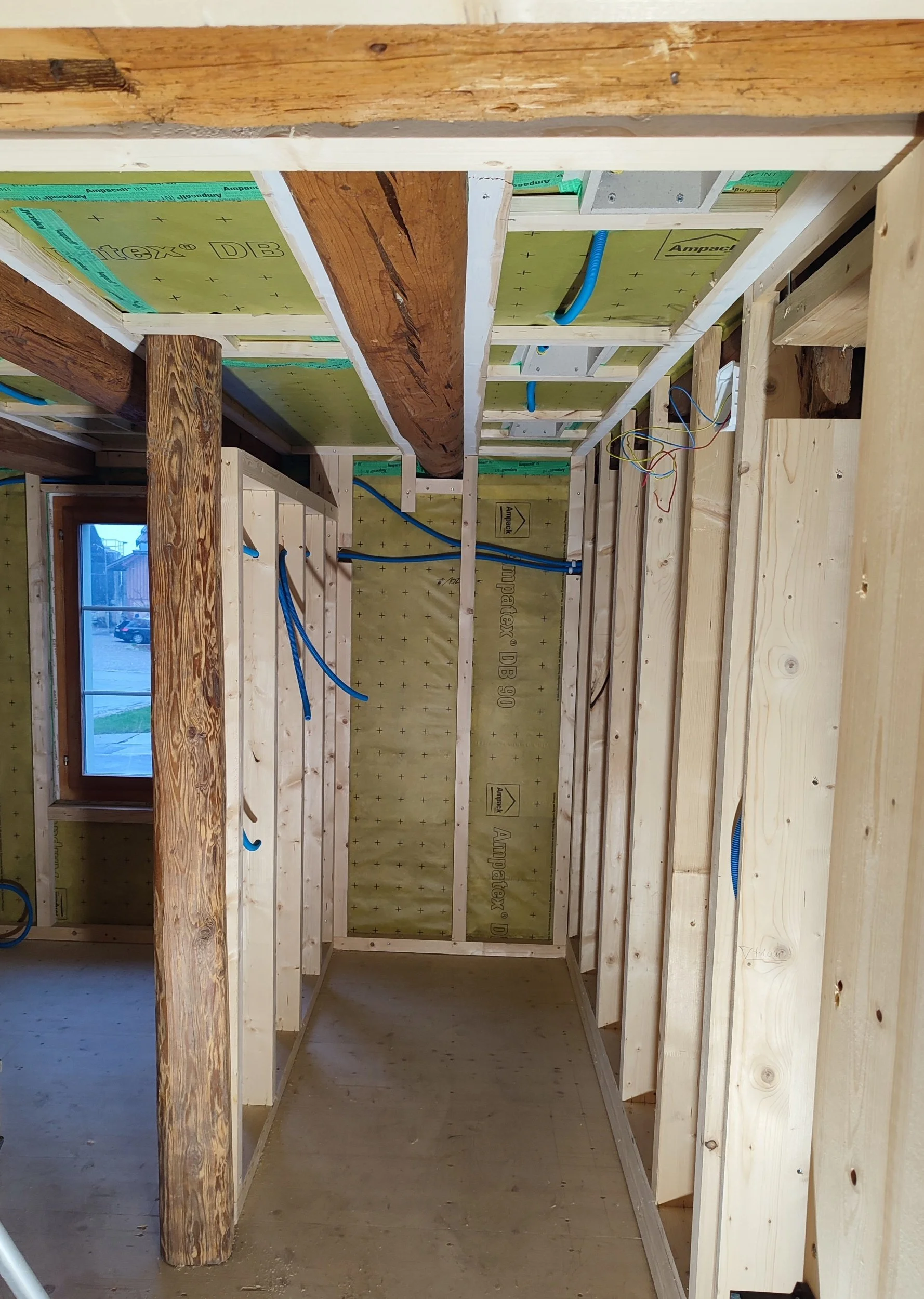 Innenansicht eines Bauprojekts, mit Holzwänden, sichtbaren elektrischen Leitungen und Isolierung, Fenster sichtbar.