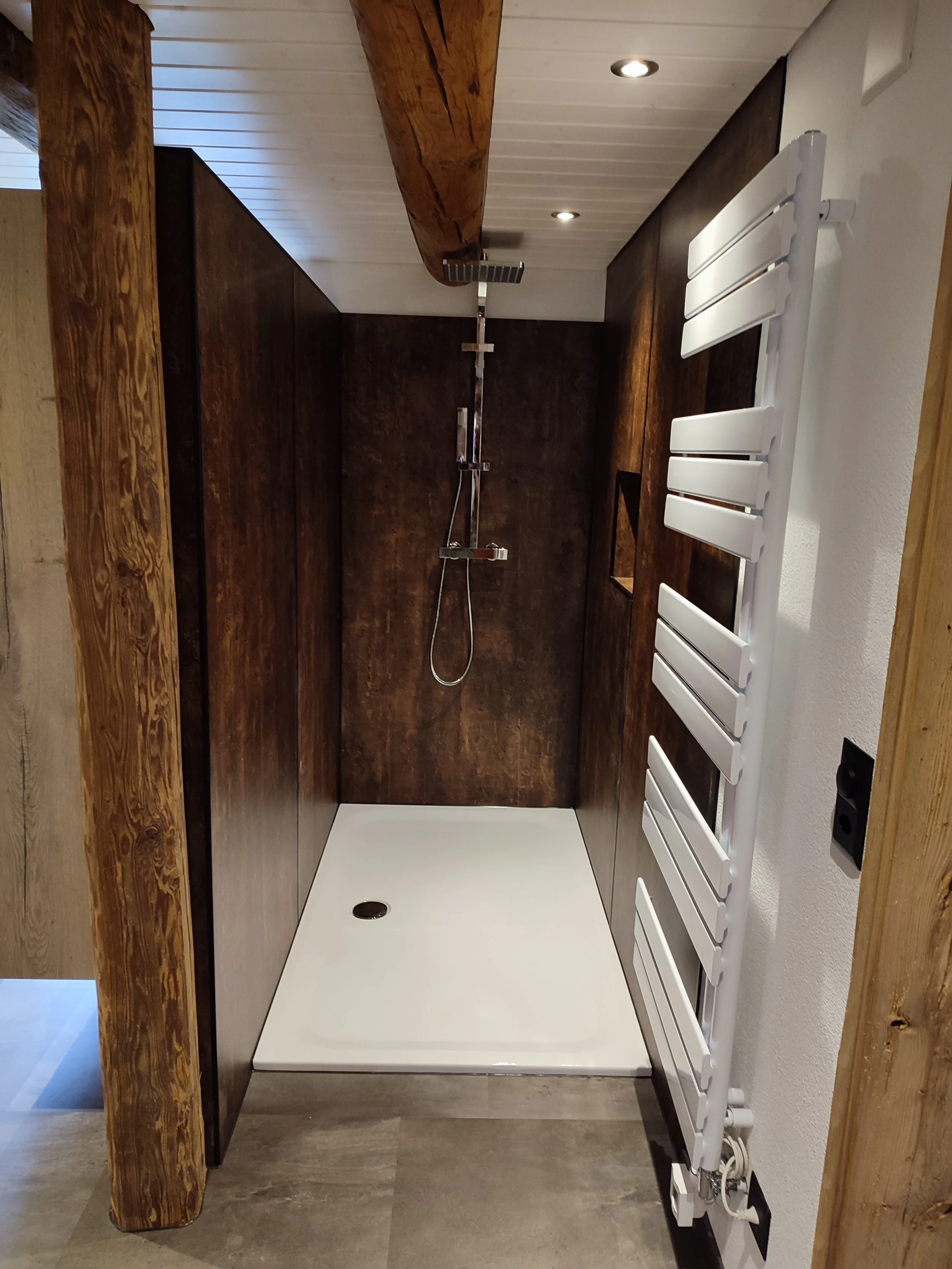 Ein minimalistisches Duschbad mit dunklen Holzwänden, einer modernen Regenwalddusche, einem weißen Boden und einem weißen Heizkörper an der rechten Wand.