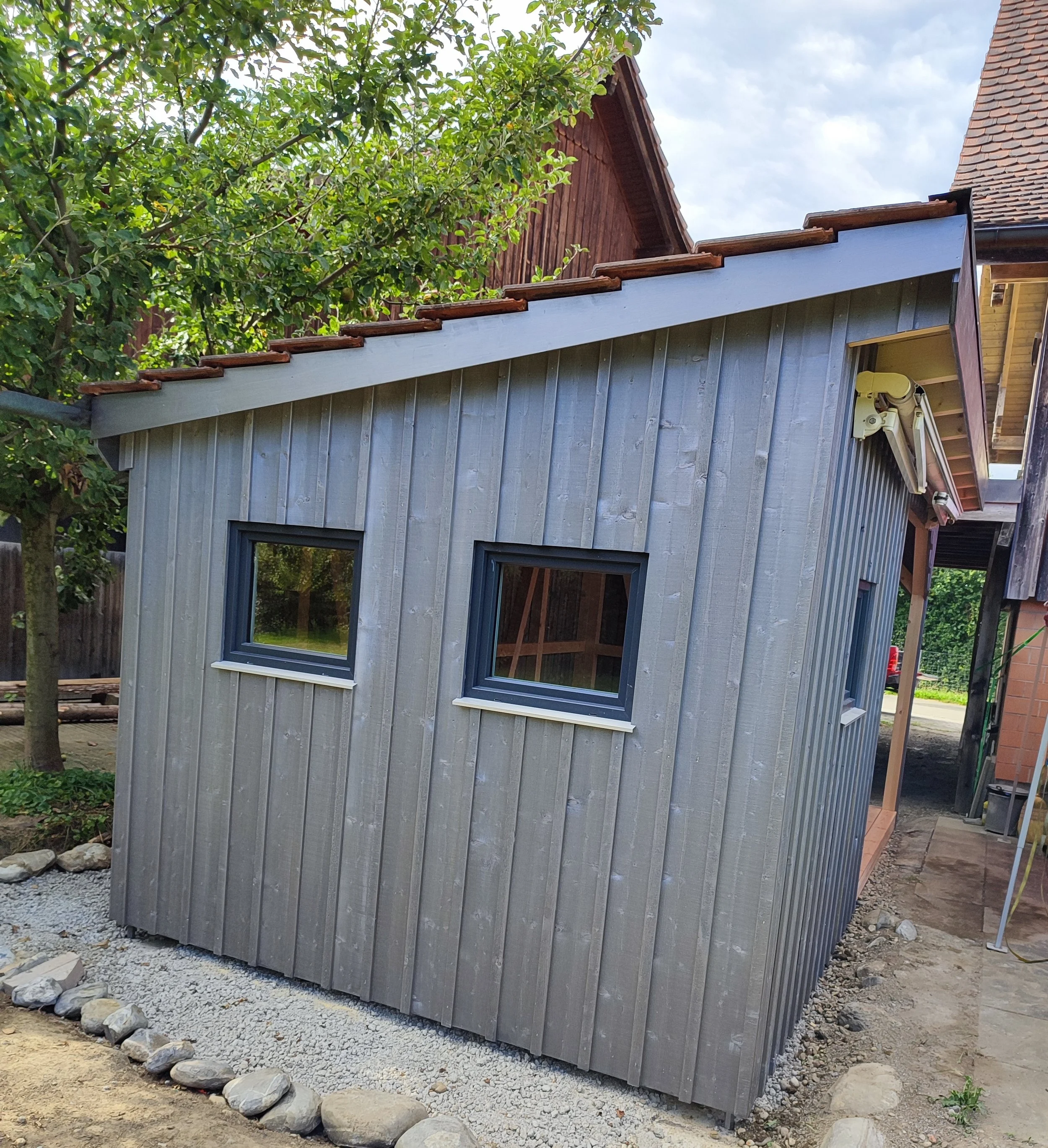 Kleines Gartenhaus mit silberner Vertikaltasung und zwei kleine Fenster, umgeben von einer Kiesumrandung und Bäumen im Hintergrund.