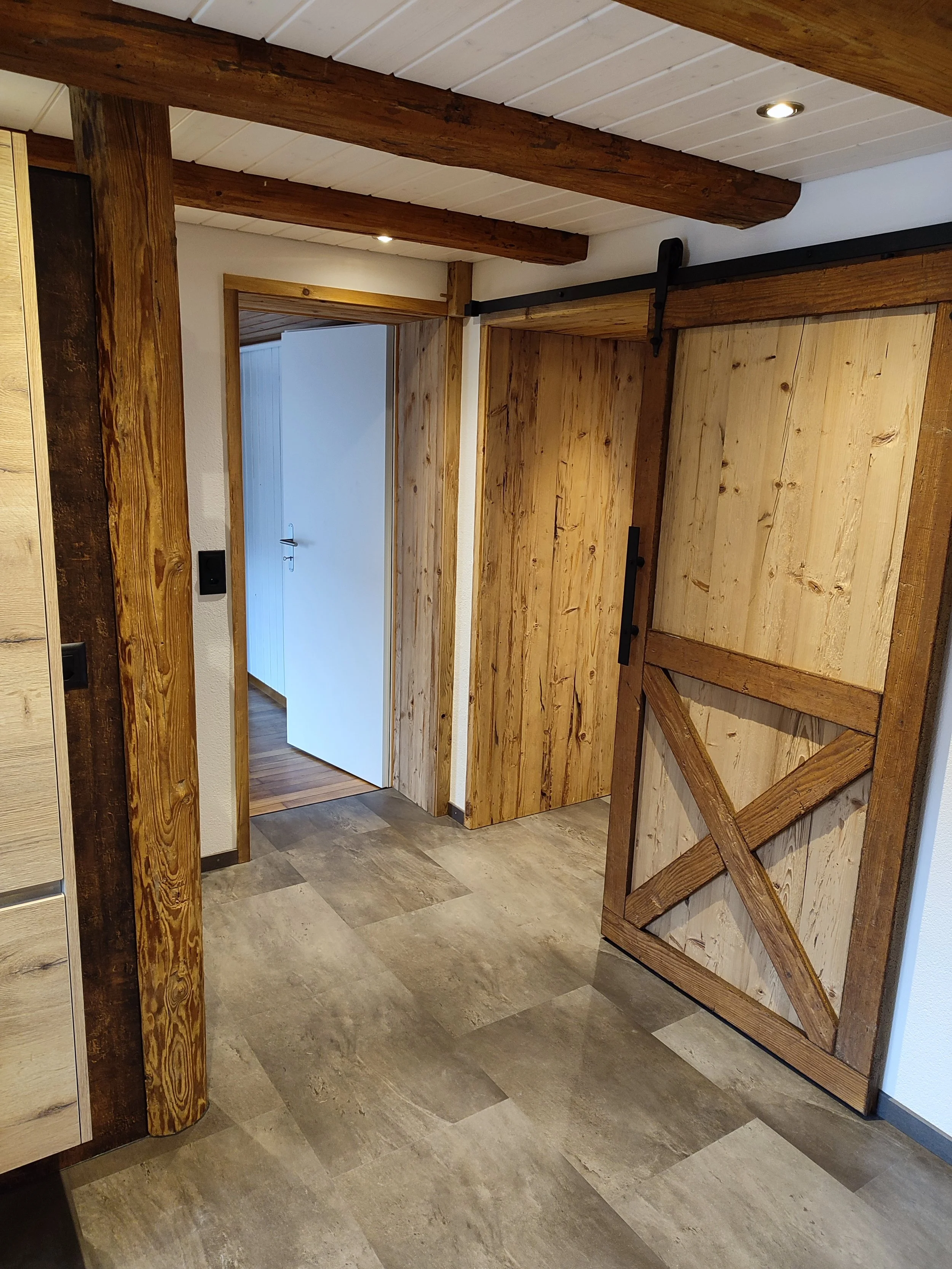 Innenraum mit Holztafeln und -rahmen, einer Schiebetür auf Schiene, modernem Fliesenboden, weißer Wandfarbe und Holzbalken an der Decke.