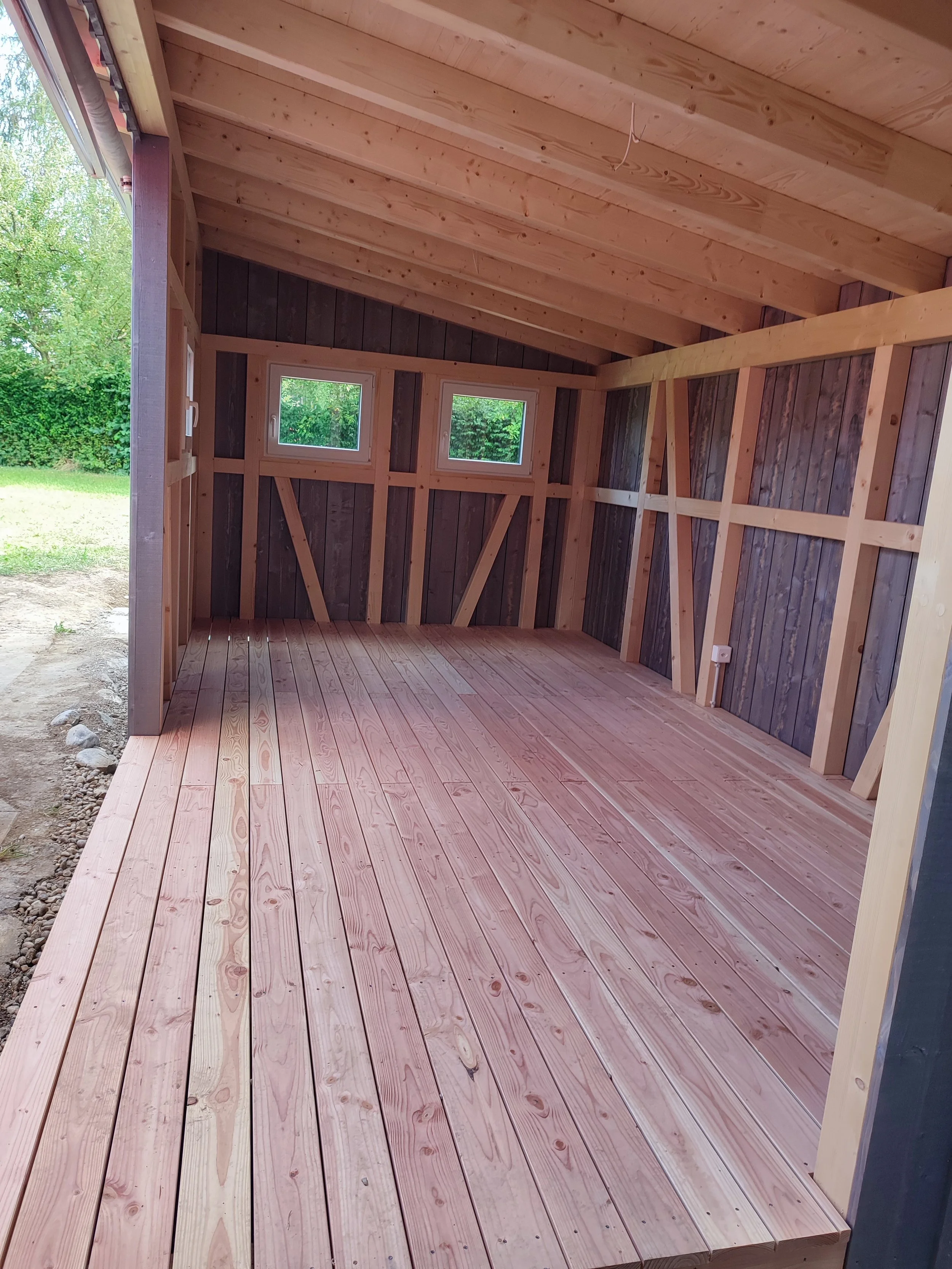Innenraum eines unvollendeten Holzschuppens mit Holzboden, Wänden und Dachkonstruktion, zwei kleine Fenster an der Rückwand