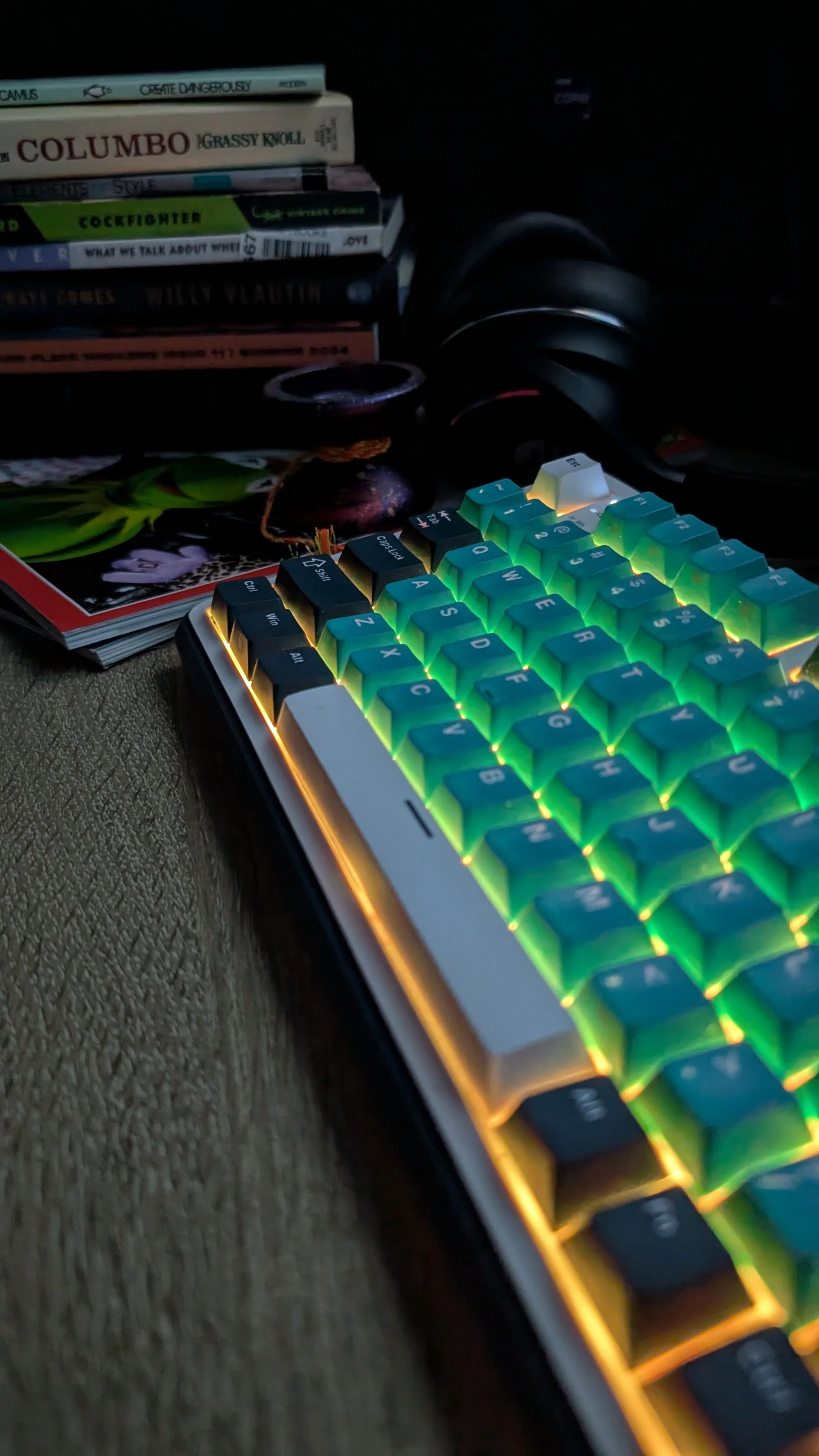 A close-up of a colorful, illuminated mechanical keyboard on a textured surface, with a stack of books and a magazine in the background.