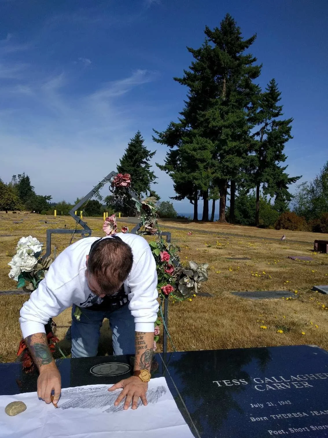 Man with tattoos leaning over a grave, drawing on a white cloth in a cemetery with large trees in the background.