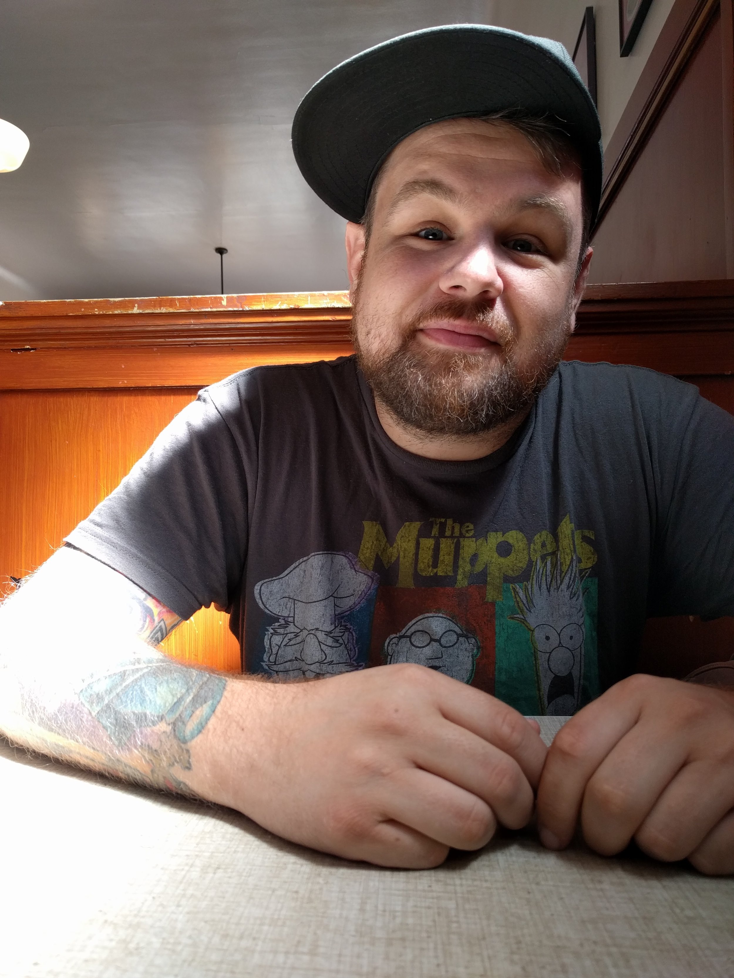 A man with a beard and tattoos is wearing a black cap backwards and a The Muppets T-shirt. He is sitting at a table in a restaurant or cafe, leaning forward with his hands clasped, and looking at the camera with a slight smile.