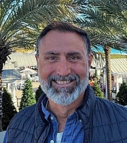 Man smiling outdoors with palm trees and buildings in the background.